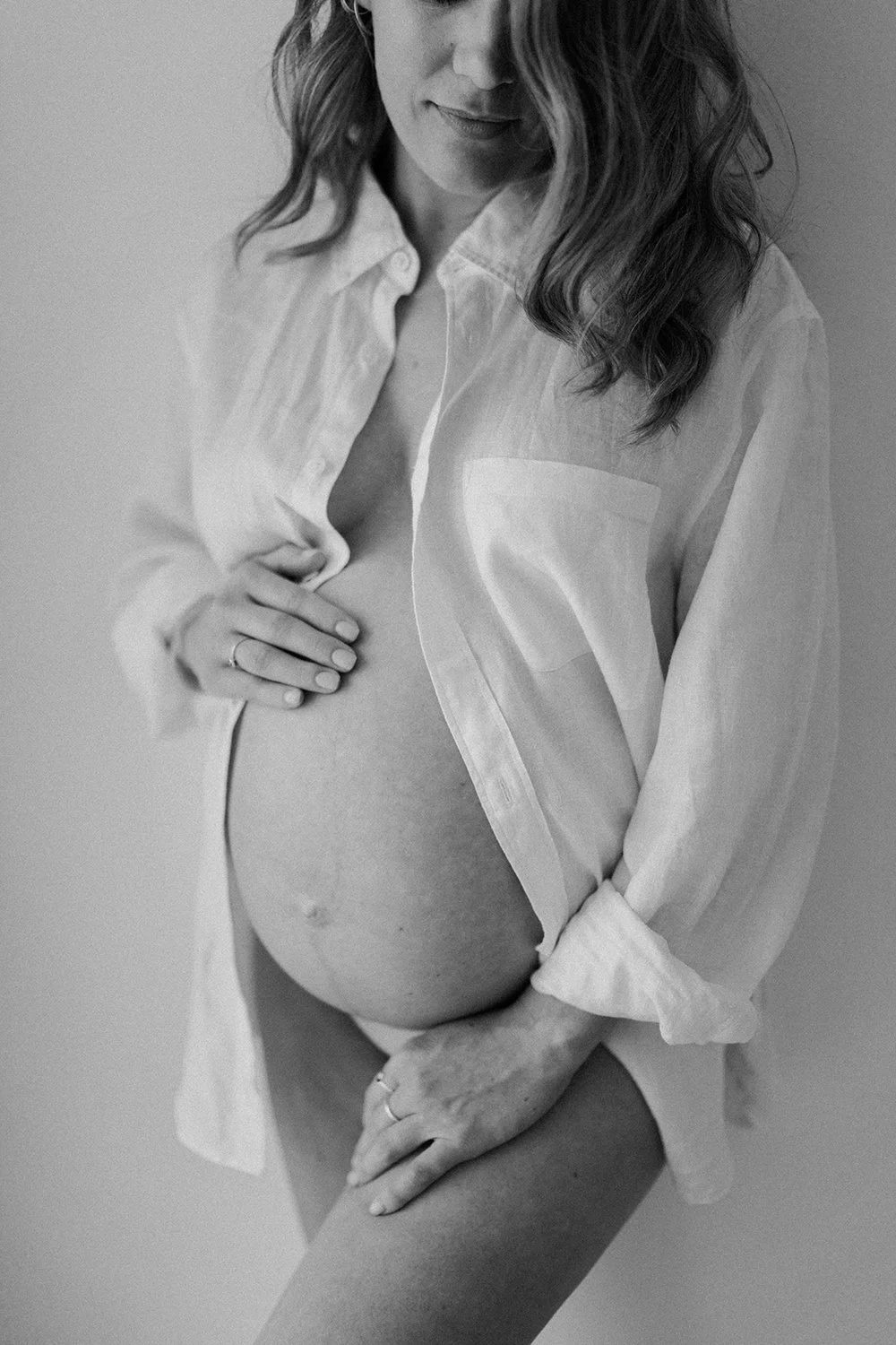 A woman in a partially unbuttoned white shirt, revealing her pregnant belly, with curly hair and a gentle smile, standing against a plain background.