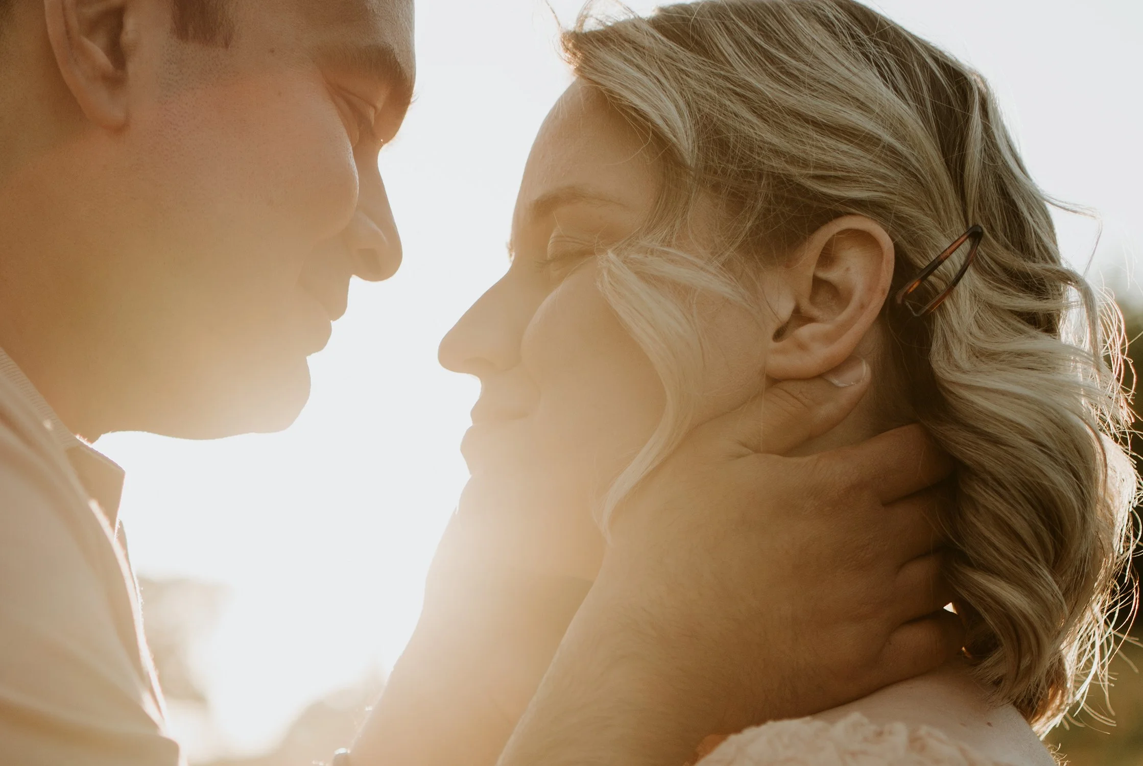 A man and a woman are very close, forehead to forehead, with the man gently holding the woman's face and the woman smiling softly. They are outdoors with bright sunlight behind them.