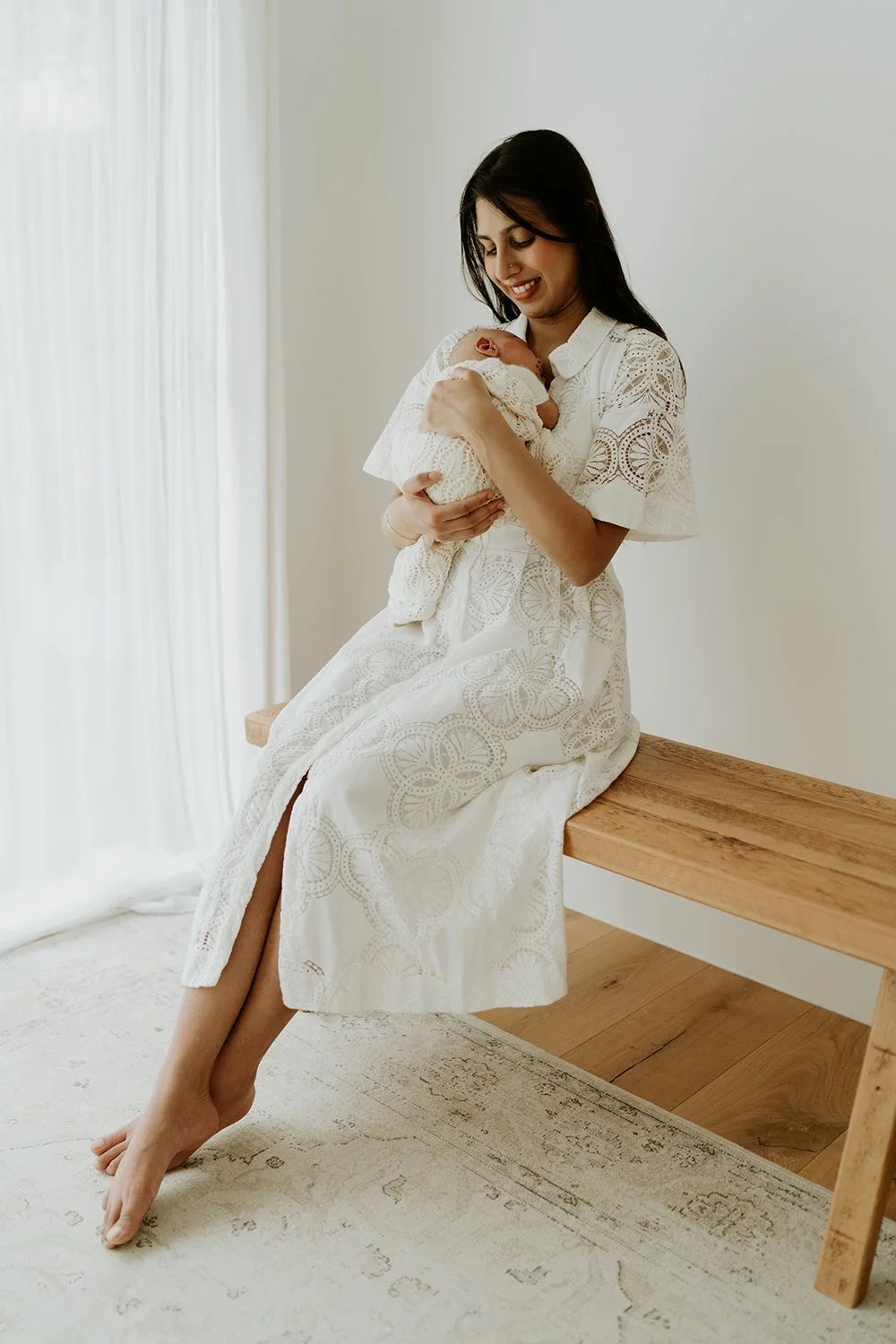 A woman sitting on a wooden bench, holding a baby close to her chest, smiling, in a bright room with white walls and curtains, dressed in a white lace dress.