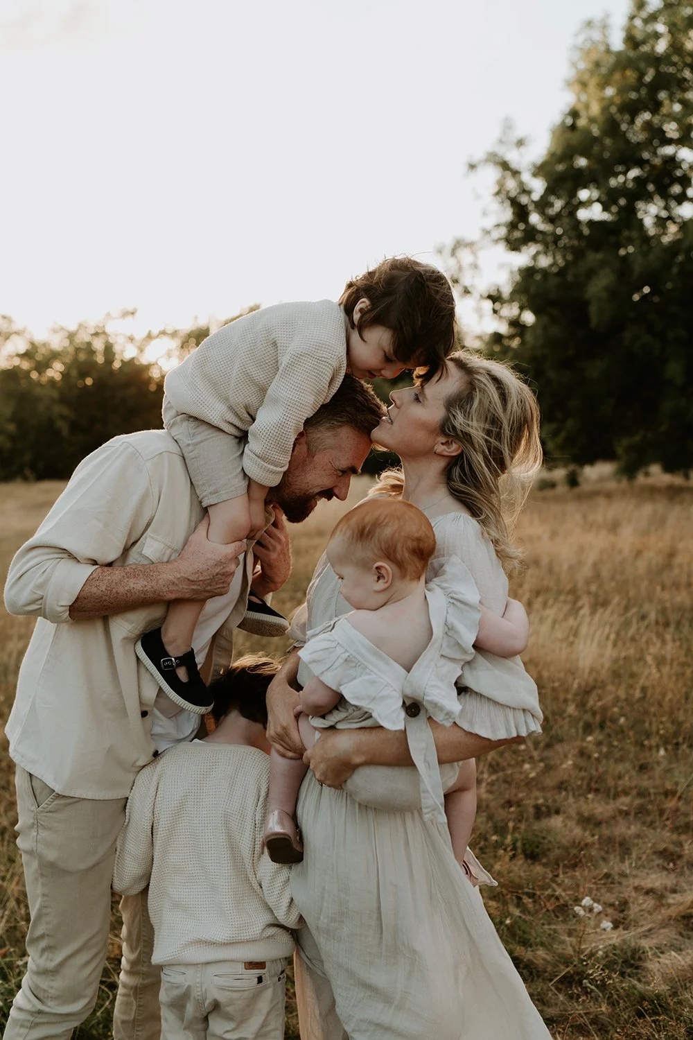 Family of five enjoying a moment outdoors at sunset, with trees in the background. The mother holds a young child, and the father carries a child on his shoulders while another child is close by.