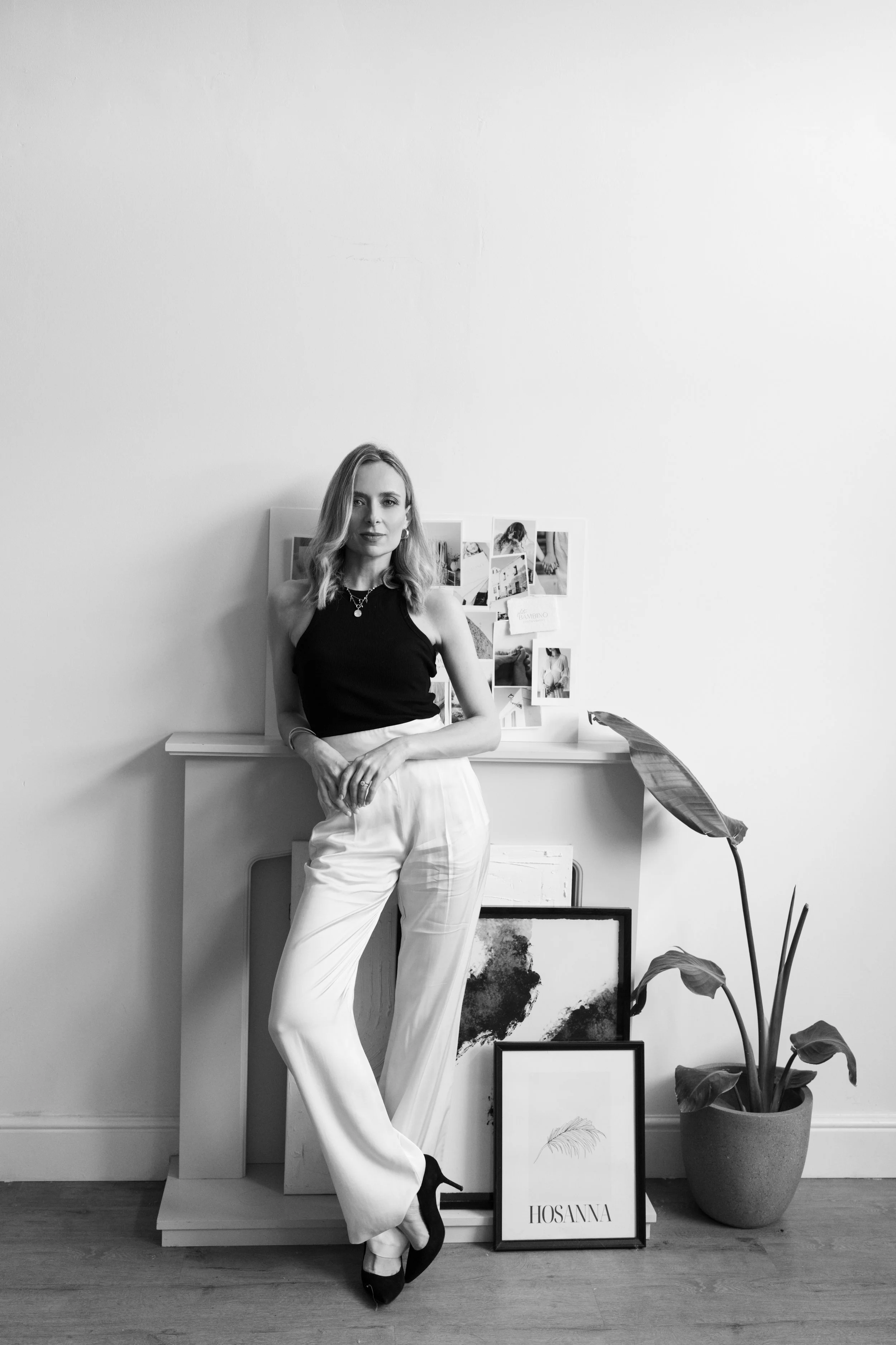 A woman with shoulder-length wavy hair, wearing a black sleeveless top and white wide-leg pants, standing in front of a white wall with framed artwork and photographs, next to a potted plant.