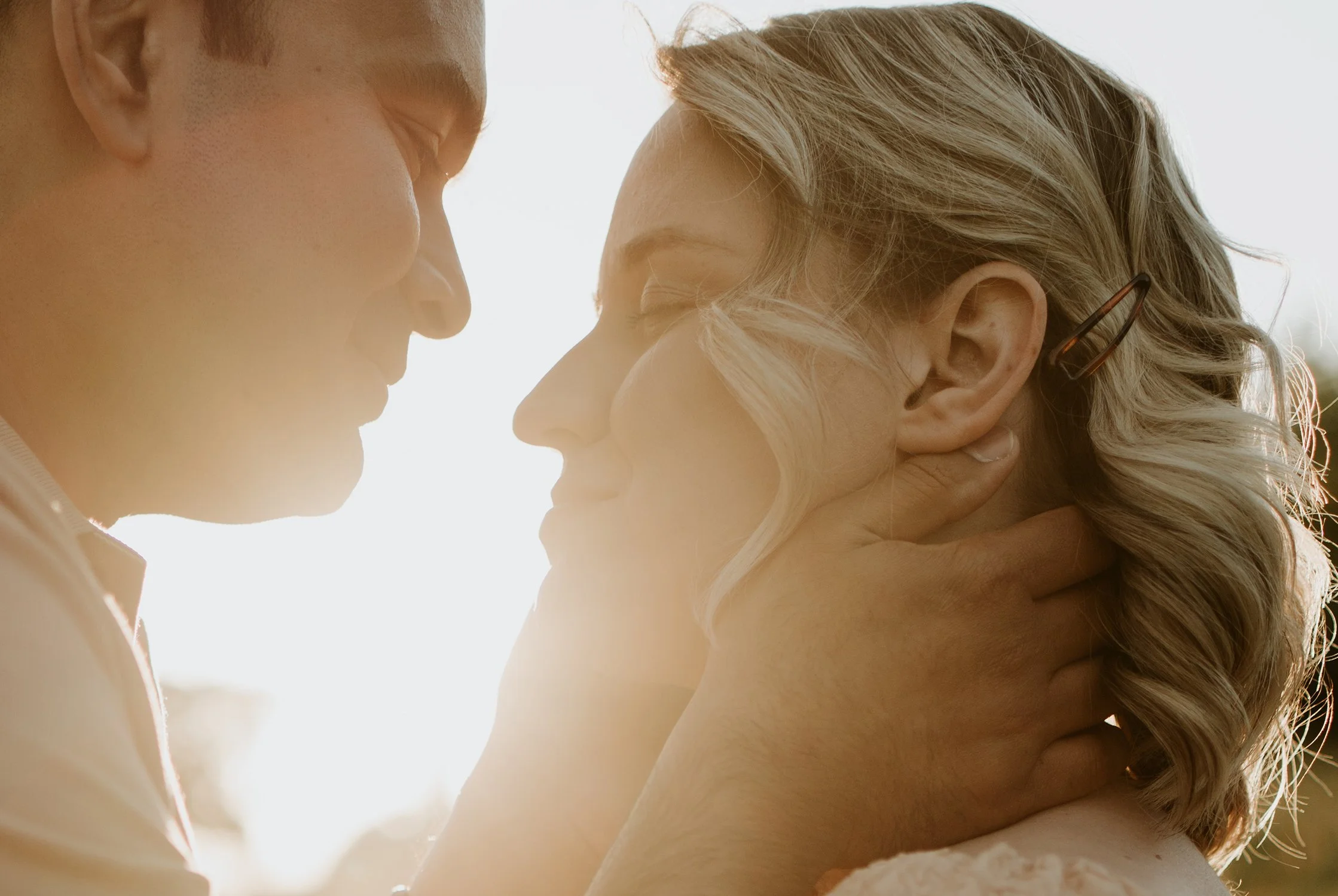 A close-up of a romantic moment between a man and a woman, with their faces gently touching and eyes closed, backlit by sunlight.