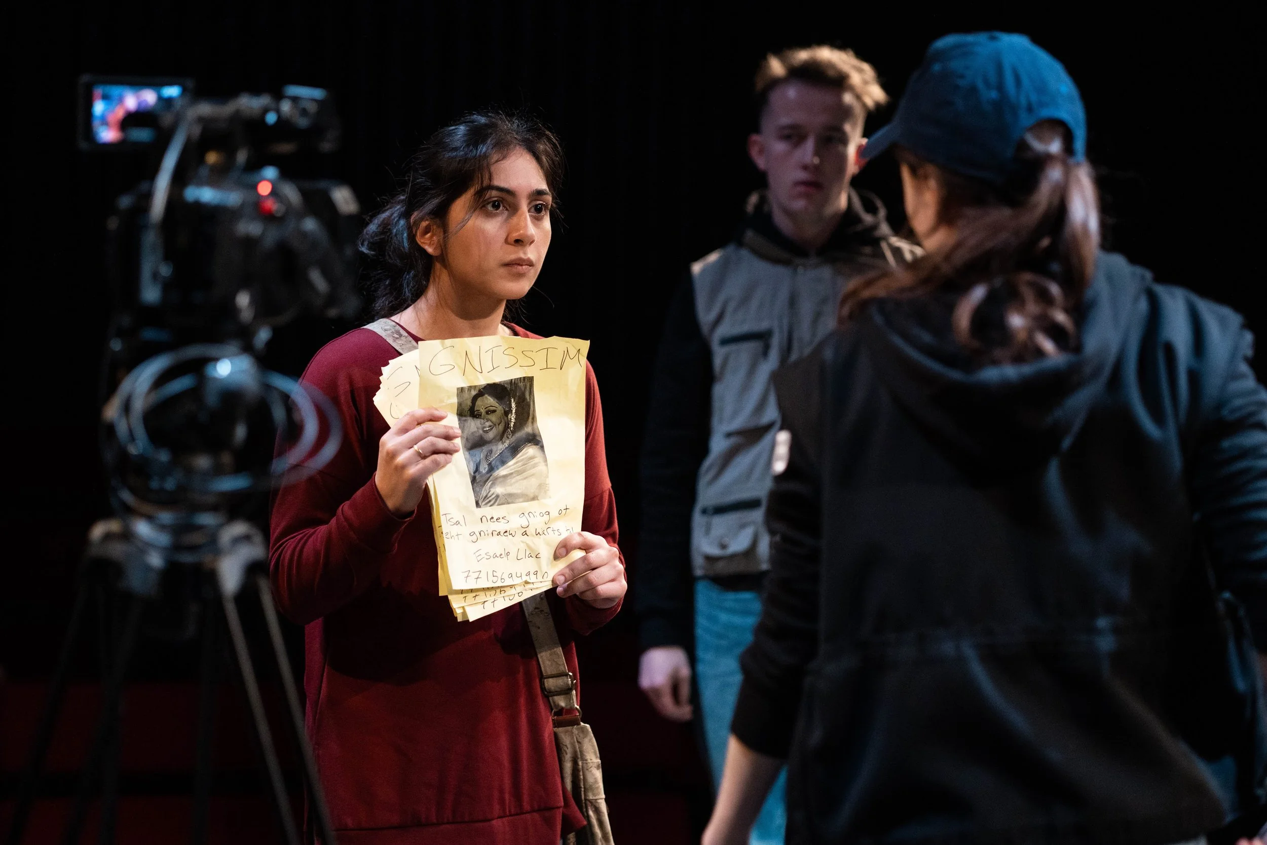 Young woman holding a handwritten poster with a photo, standing in front of a film camera during a filming scene.