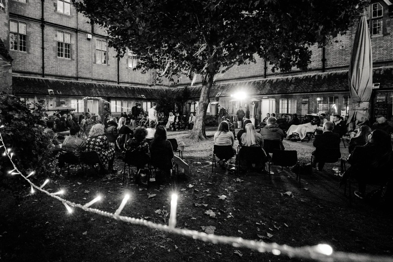 People gathered outdoors in a courtyard at night, sitting on chairs in a circle around a tree, participating in a nighttime event or gathering, with a bright light illuminating the scene and string lights visible on the left.
