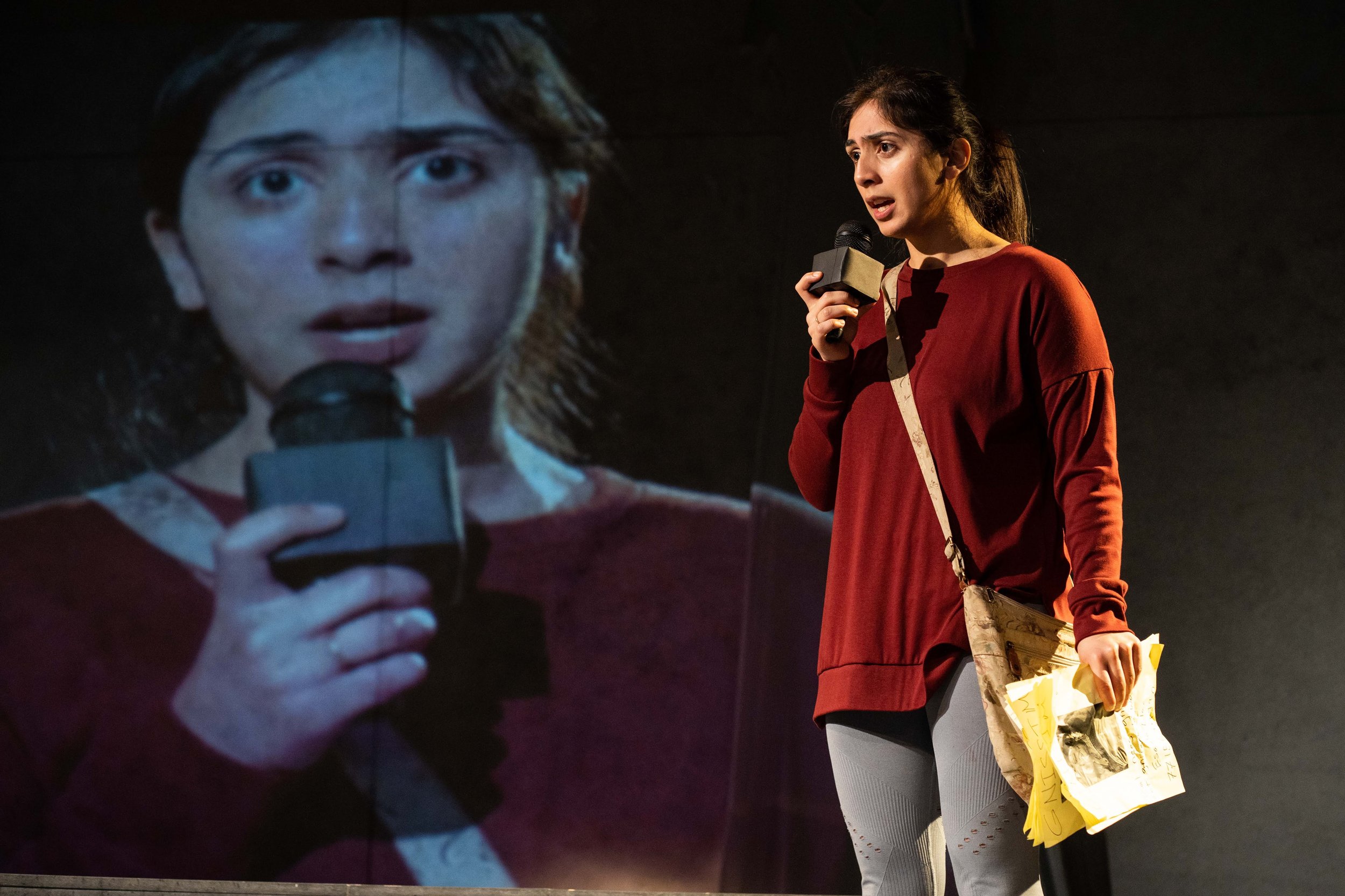 A woman in a red sweater holding a microphone and paper, speaking in front of a large screen displaying her face holding a microphone.