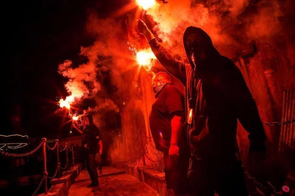 People holding lit flares amid a backdrop of flames and smoke during a nighttime protest or demonstration.