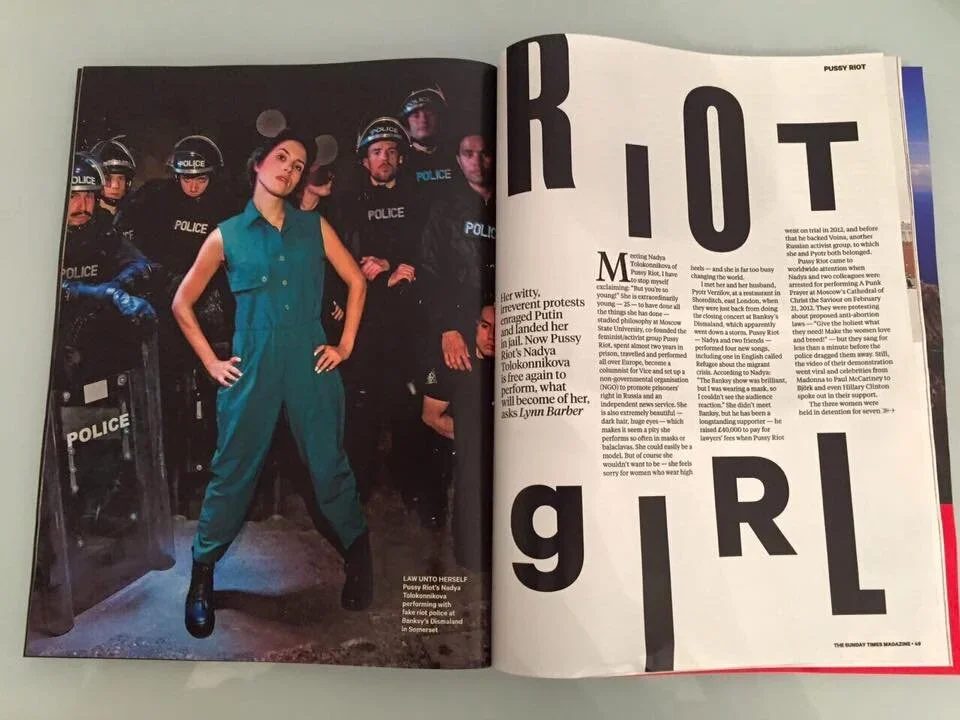 Magazine spread featuring a woman in a teal jumpsuit standing confidently with her hands on her hips surrounded by police officers in riot gear.