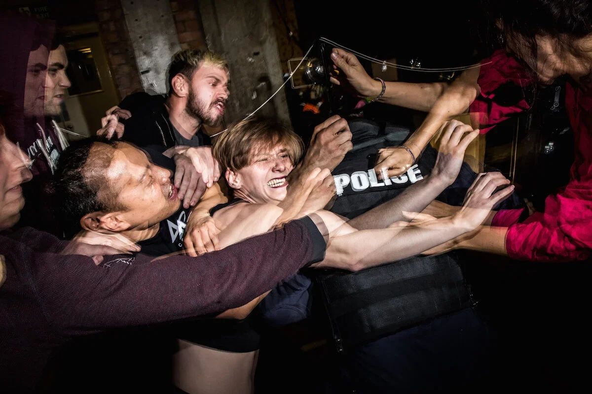 A chaotic scene of multiple people, including police officers in riot gear, forcibly restraining a woman in a confrontation. The woman appears distressed, and people around her are reaching out, trying to intervene.