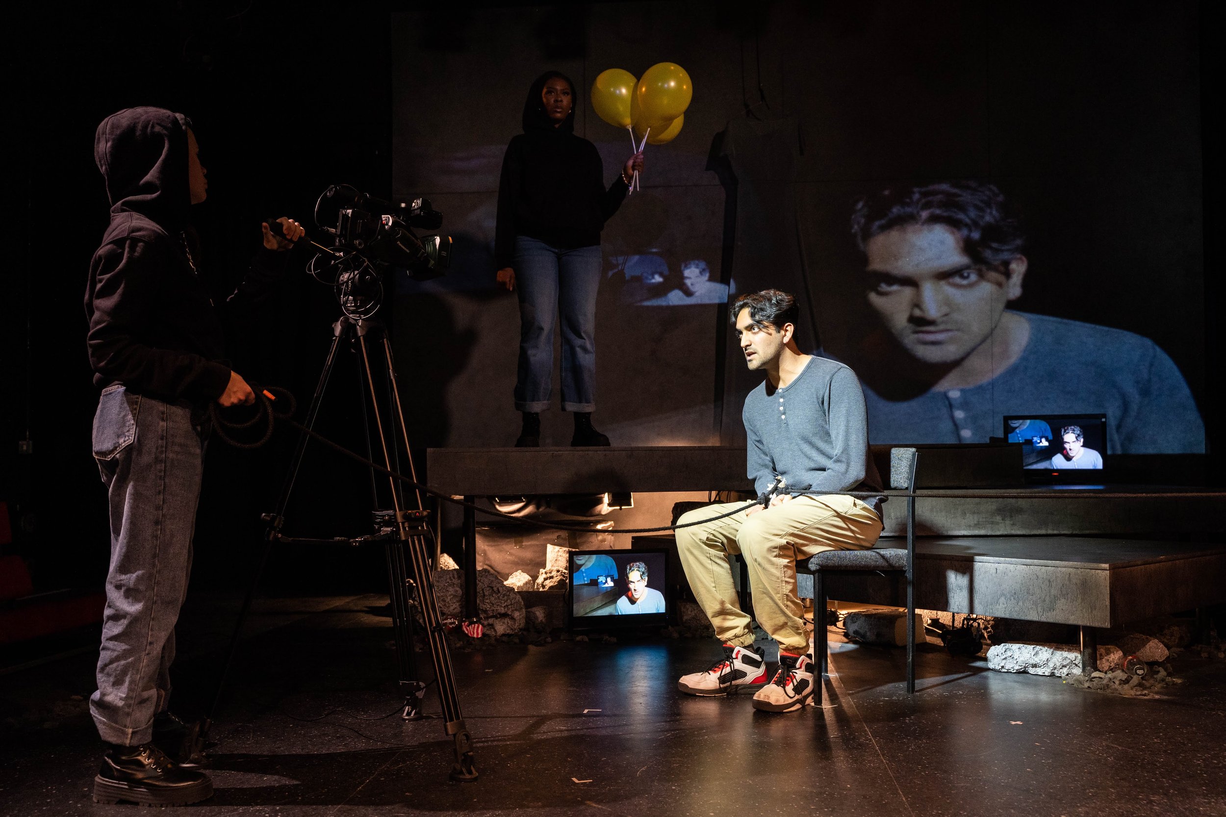 A man sitting on a bench during a film shoot with a large screen behind him displaying his face, a woman holding yellow balloons standing on a platform, and a crew member operating a camera.