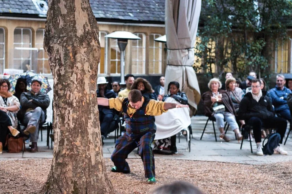 A performer in colorful plaid pants and a yellow shirt dances near a large tree in front of an audience seated outdoors. The audience watches attentively, some with umbrellas, in a courtyard with old-style buildings and trees.