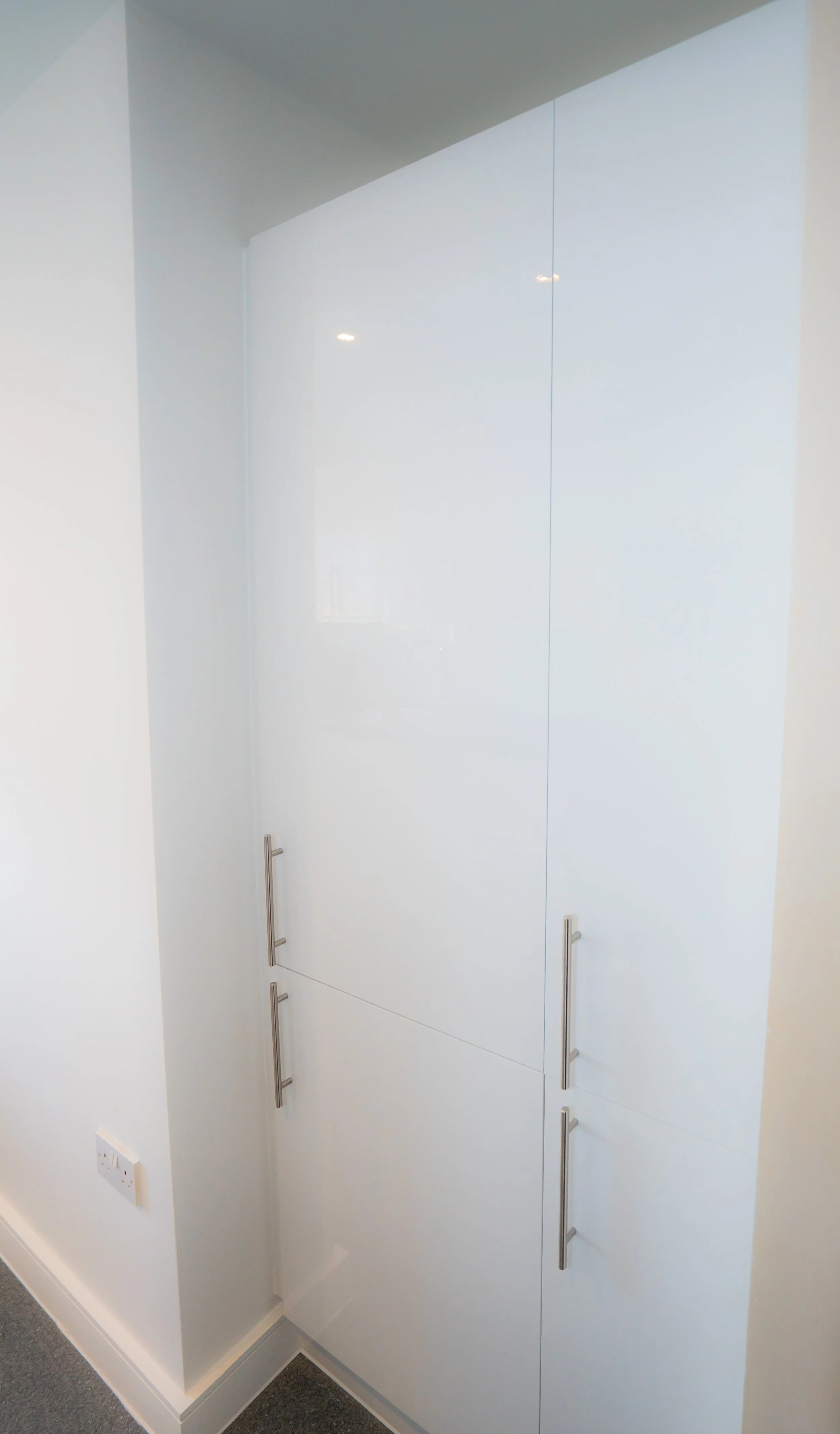 White cabinet with three vertical silver handles, mounted on a white wall in a room with a gray carpet and white baseboards, adjacent to a white wall with a power outlet.