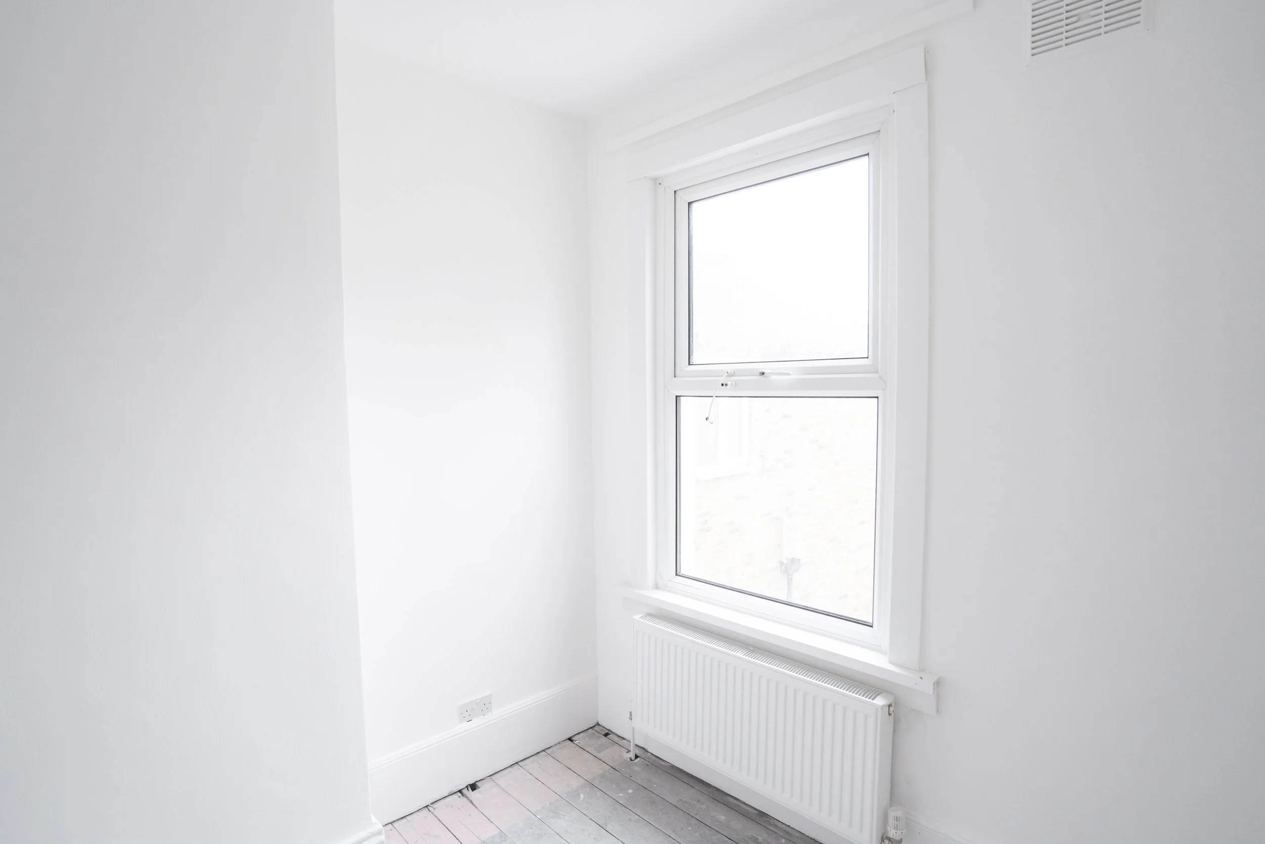 A bright room with white walls and a white door with a glass window. There is a radiator below the window and visible electrical outlets near the floor. The floor appears to be unfinished wood.