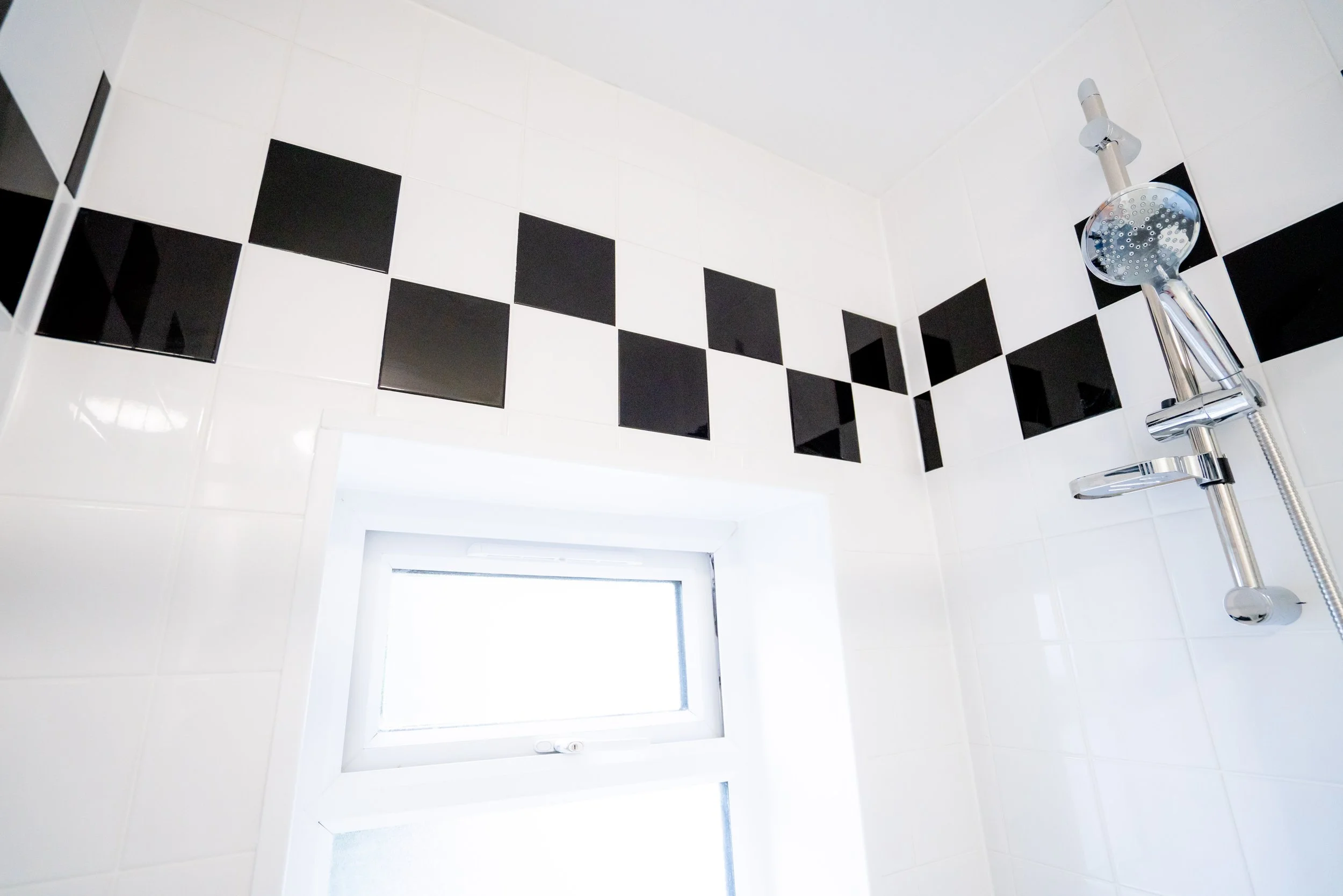 A bathroom shower with white tiles and black square tiles arranged in a line near the ceiling. There is a round showerhead attached to a vertical chrome pipe and a handheld showerhead hanging on the side. A small window is seen at the lower part of t