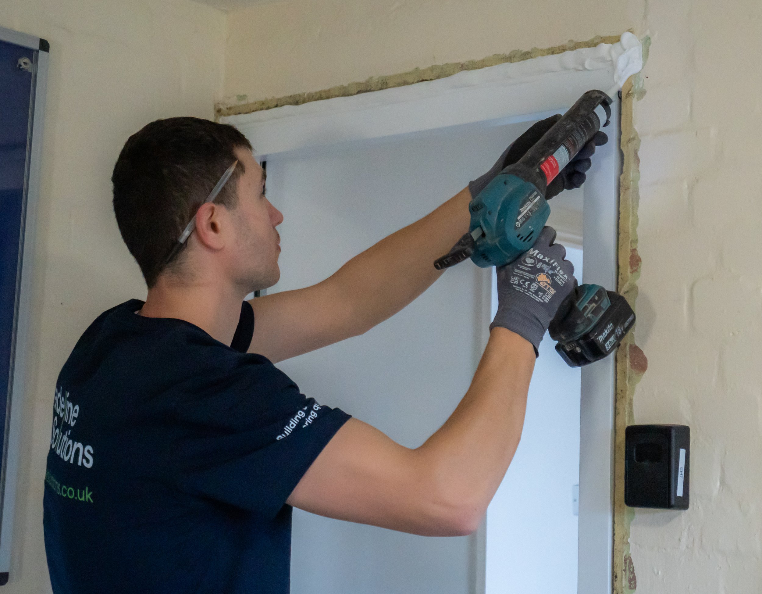 A person working on the top of a doorway frame with construction tools. They are wearing gloves, a dark T-shirt with text, and safety glasses.