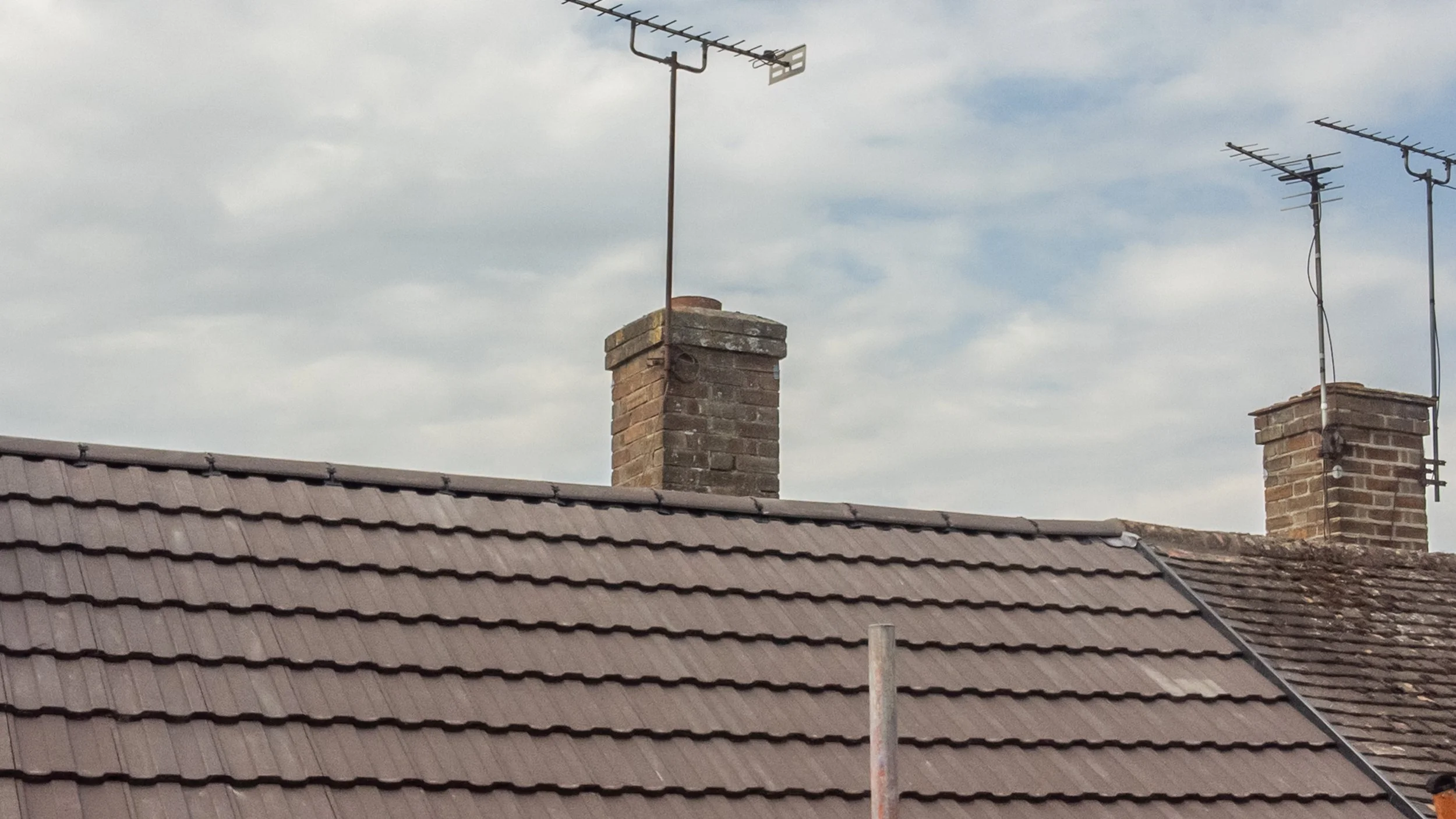 Multiple television antennas on brick chimneys and a tiled roof with a cloudy sky in the background.