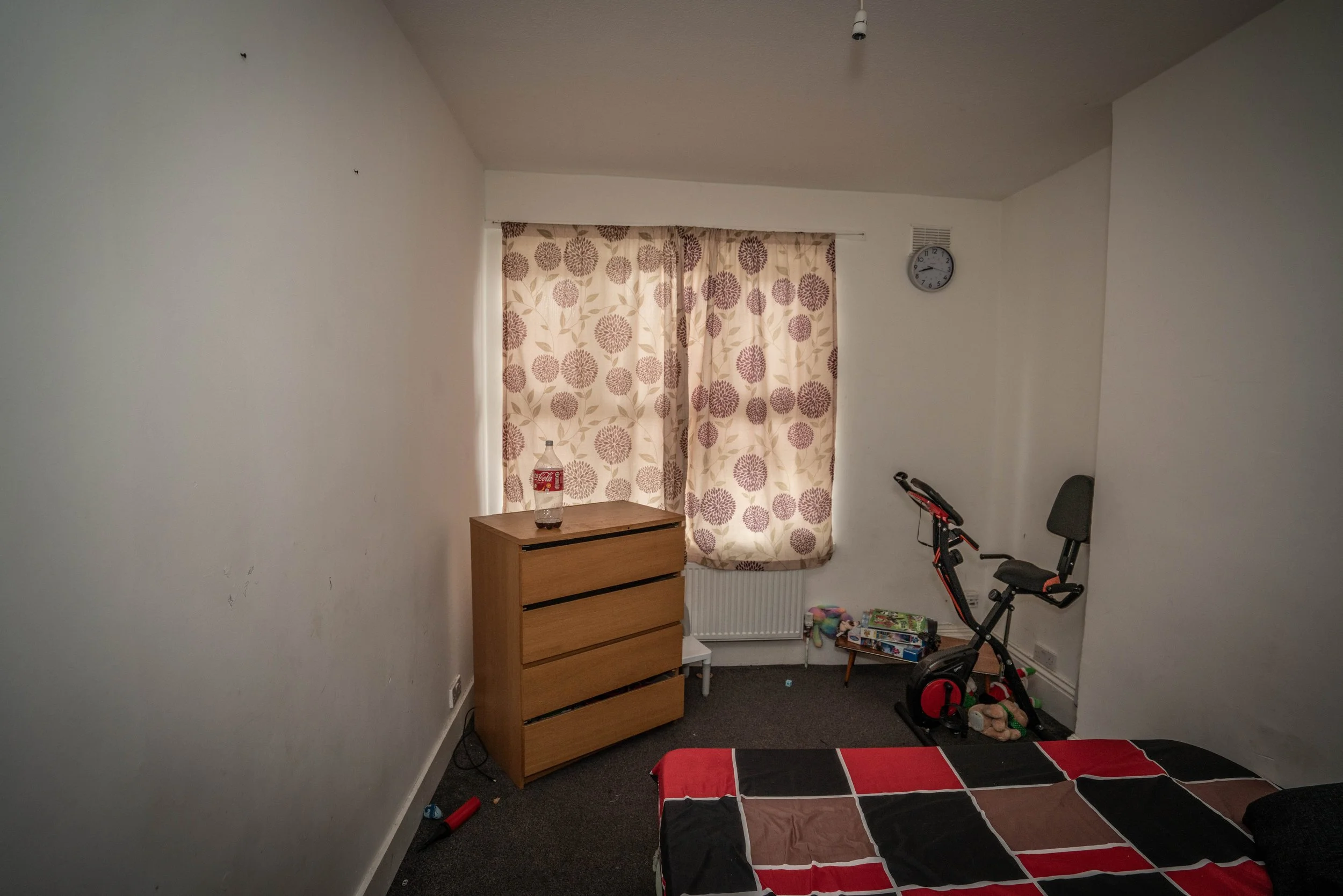 A small bedroom with off-white walls, a wooden chest of drawers, a curtain with purple floral patterns covering a window, a wall clock showing 9:08, a green gaming chair, a foldable exercise bike, a pile of toys on the floor, and a bed with a black a