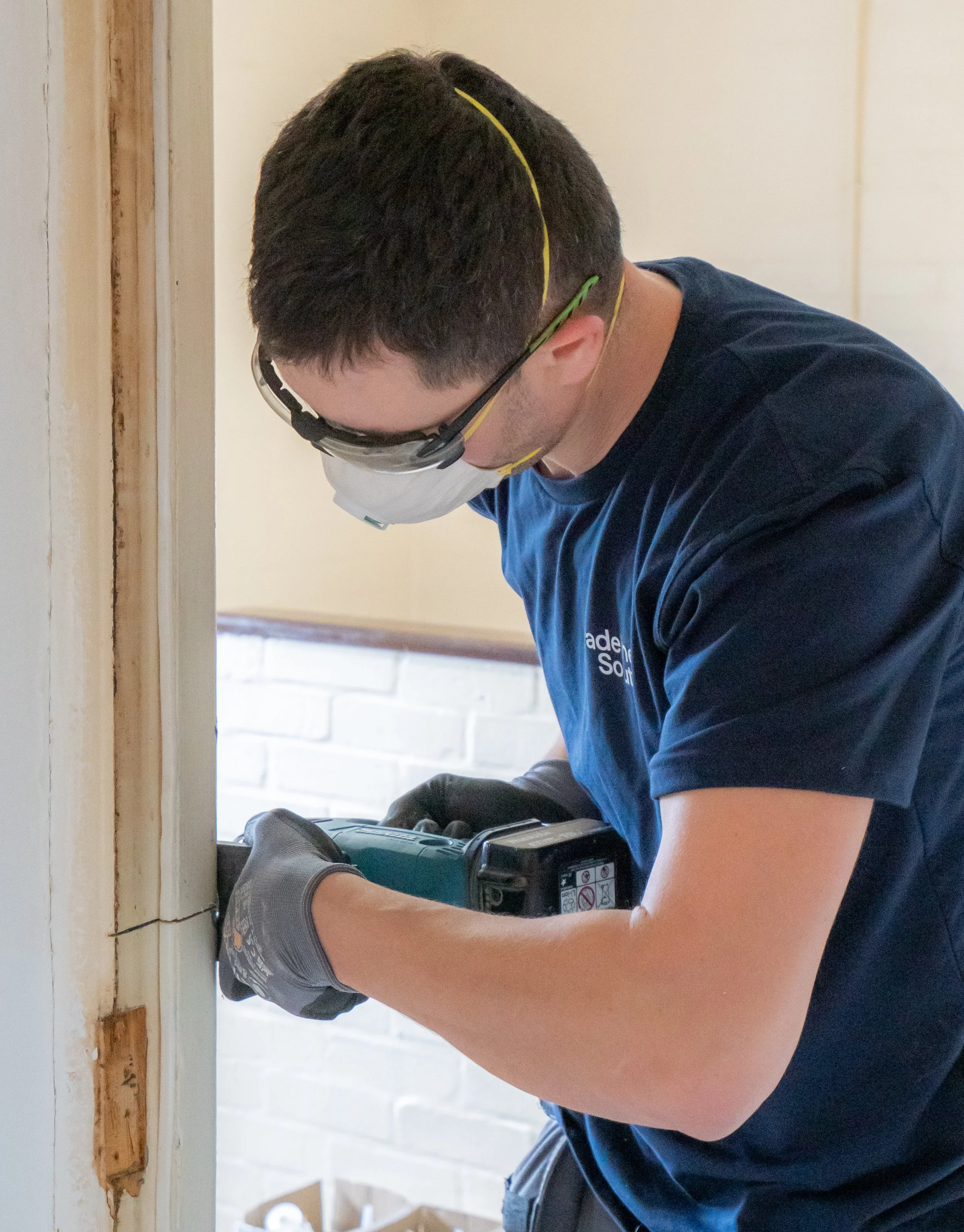 A man wearing glasses and a dust mask using a power drill to work on wood, with his arms and part of his torso visible, working indoors with a background of a white wall and a brick wall.