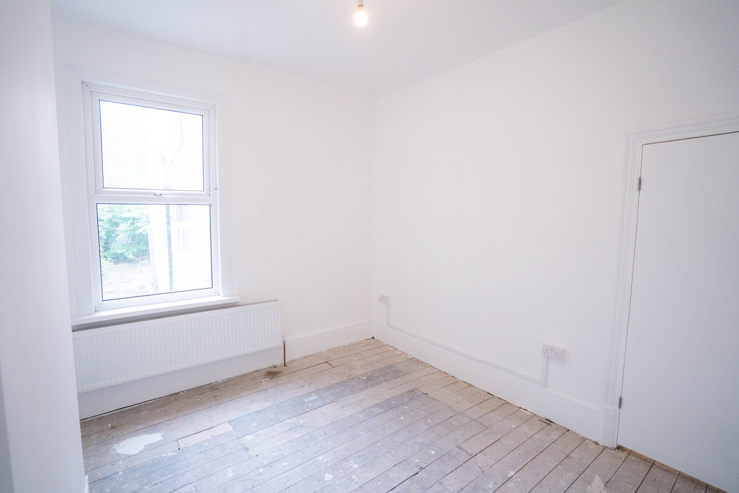 Empty white room with a large window, wooden floor, and radiator beneath the window.