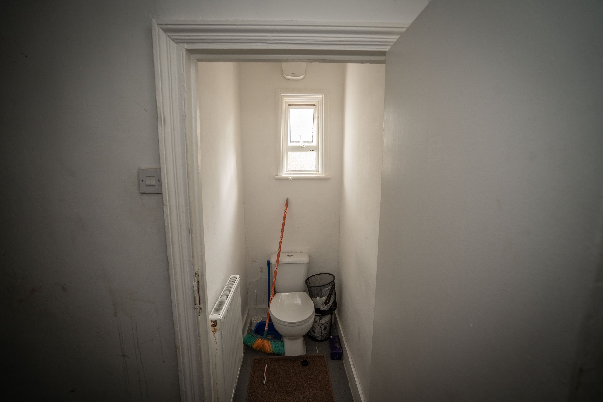 Small hallway bathroom with a toilet, a window above, and cleaning tools such as a broom and a trash bin. The walls are painted white, and there is a radiator on the left side.