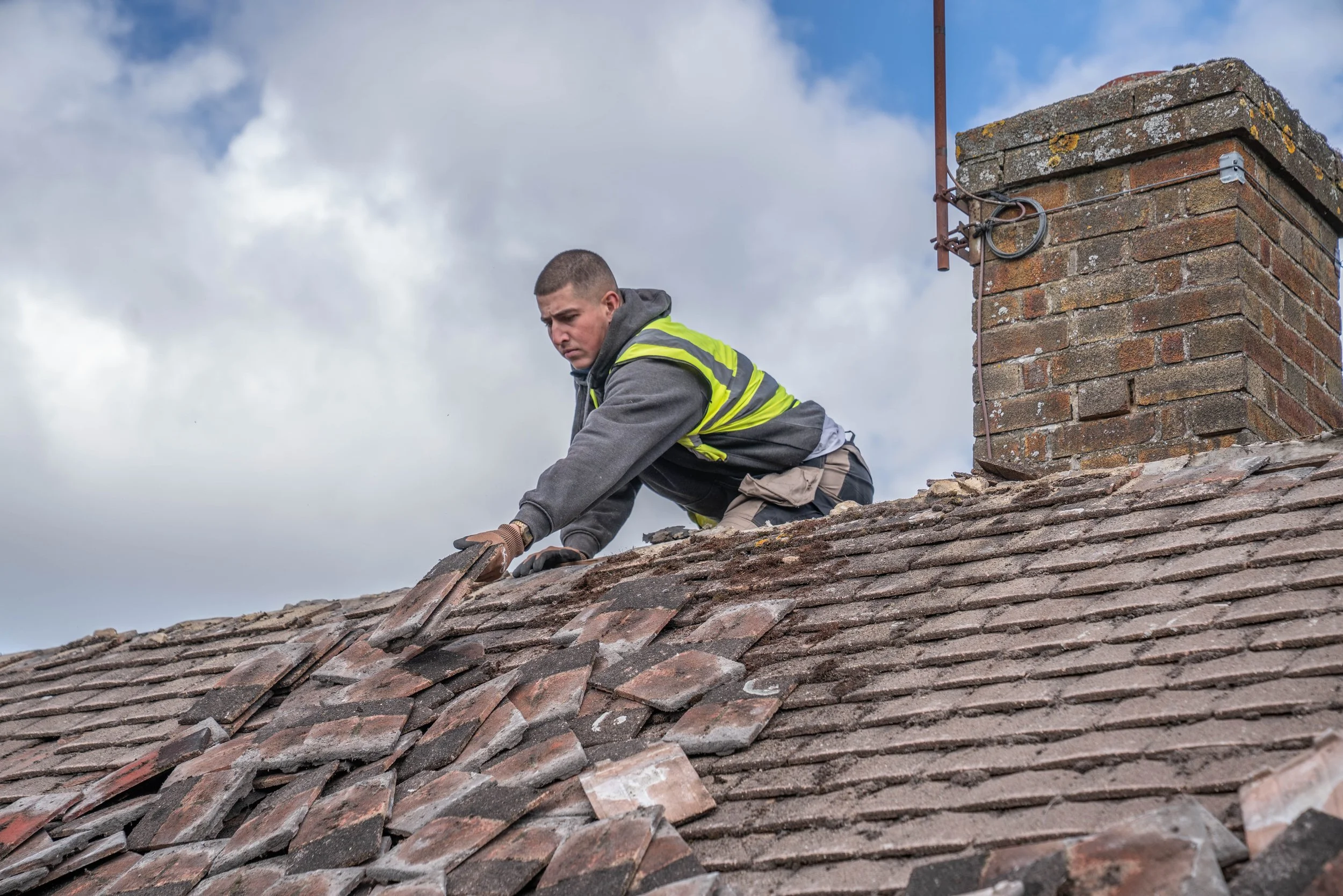 A man wearing a gray hoodie and yellow safety vest is working on a roof, repairing or inspecting the shingles beside a brick chimney, under a partly cloudy sky.