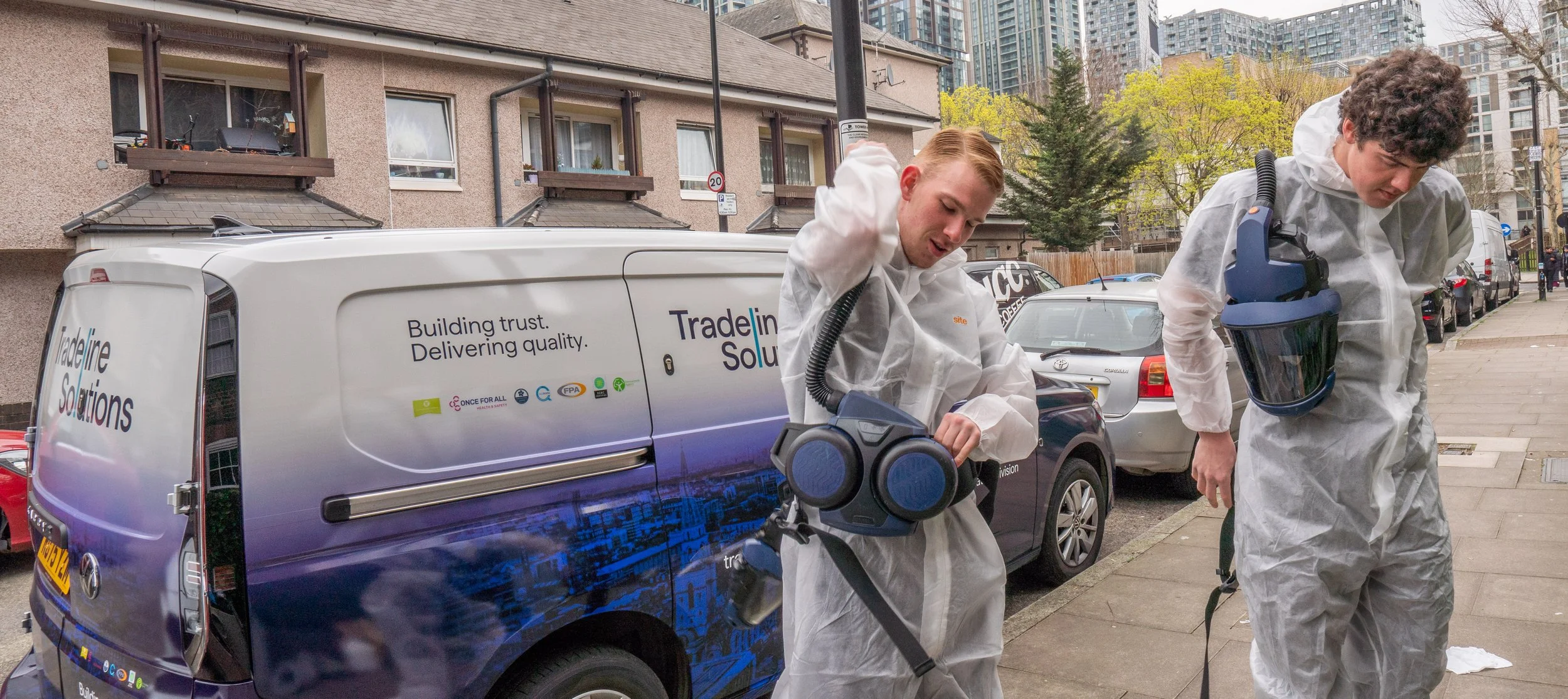 Two individuals in white protective suits amidst urban setting, with a trade solutions vehicle in the background.