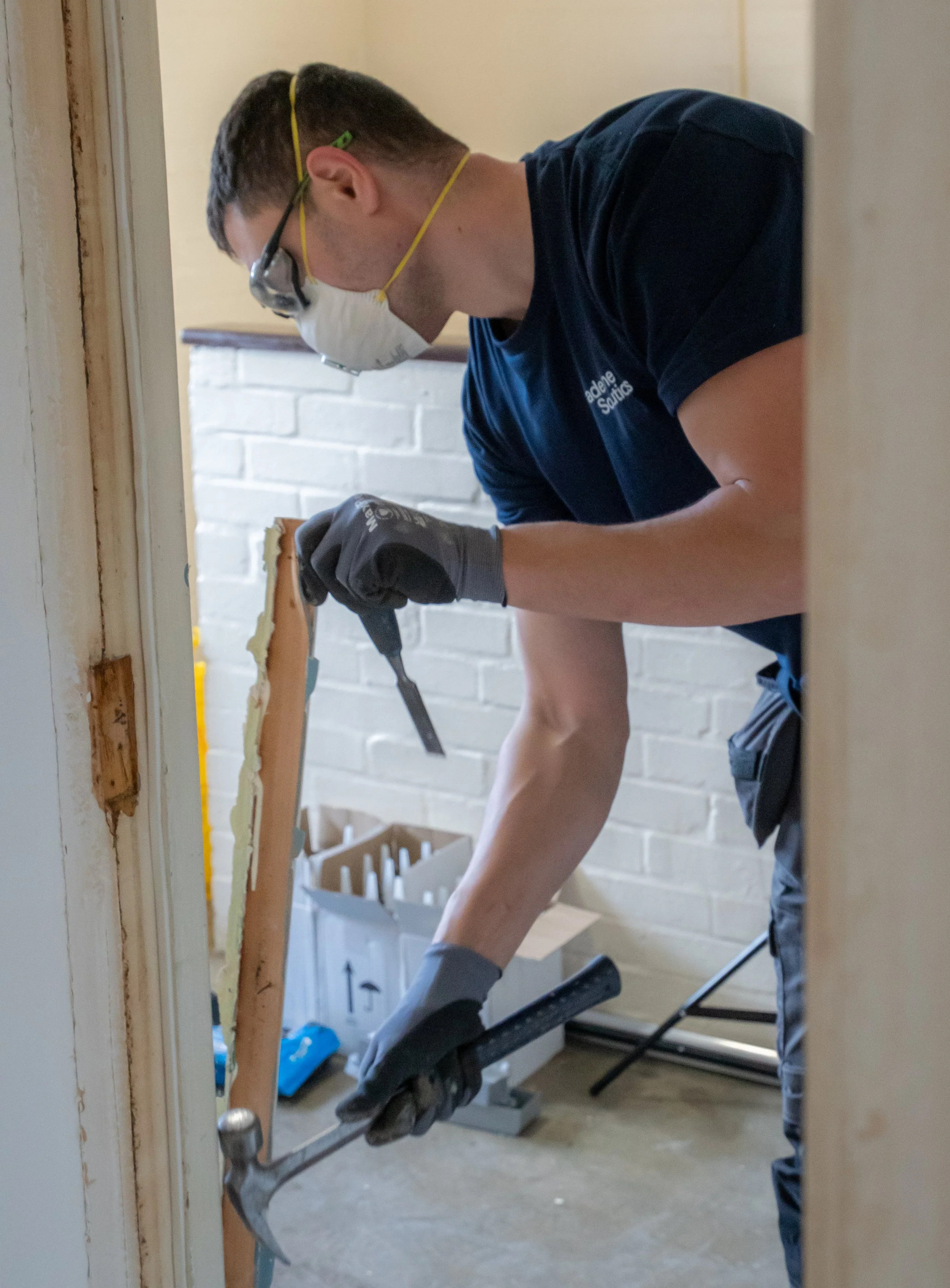 A person wearing safety goggles, a face mask, and work gloves is using a hammer and chisel to remove something from a wooden door frame while standing on a step ladder.