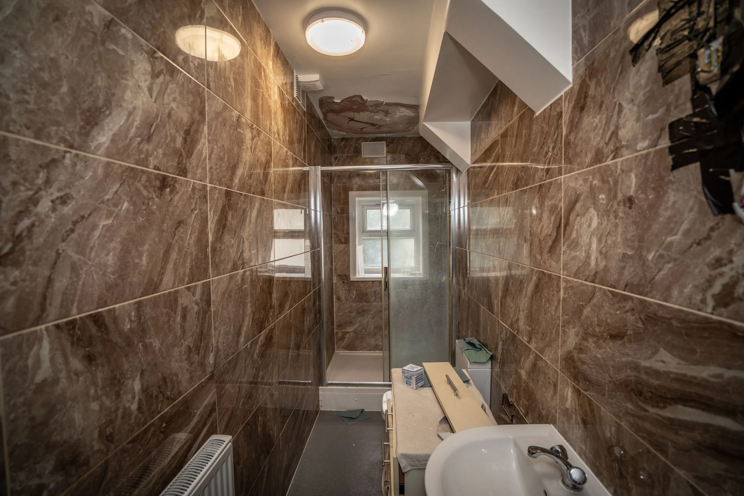 Interior view of a small bathroom with brown marble tiles, a white sink, a mirror, a small window, and various items on the counter including a box of tissues and some toiletries.
