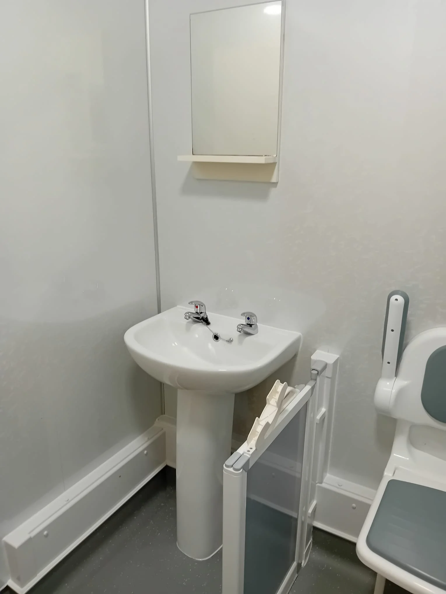 An accessible bathroom with a white sink, a mirror above it, a white foot pedal sink, and a wall-mounted toilet.