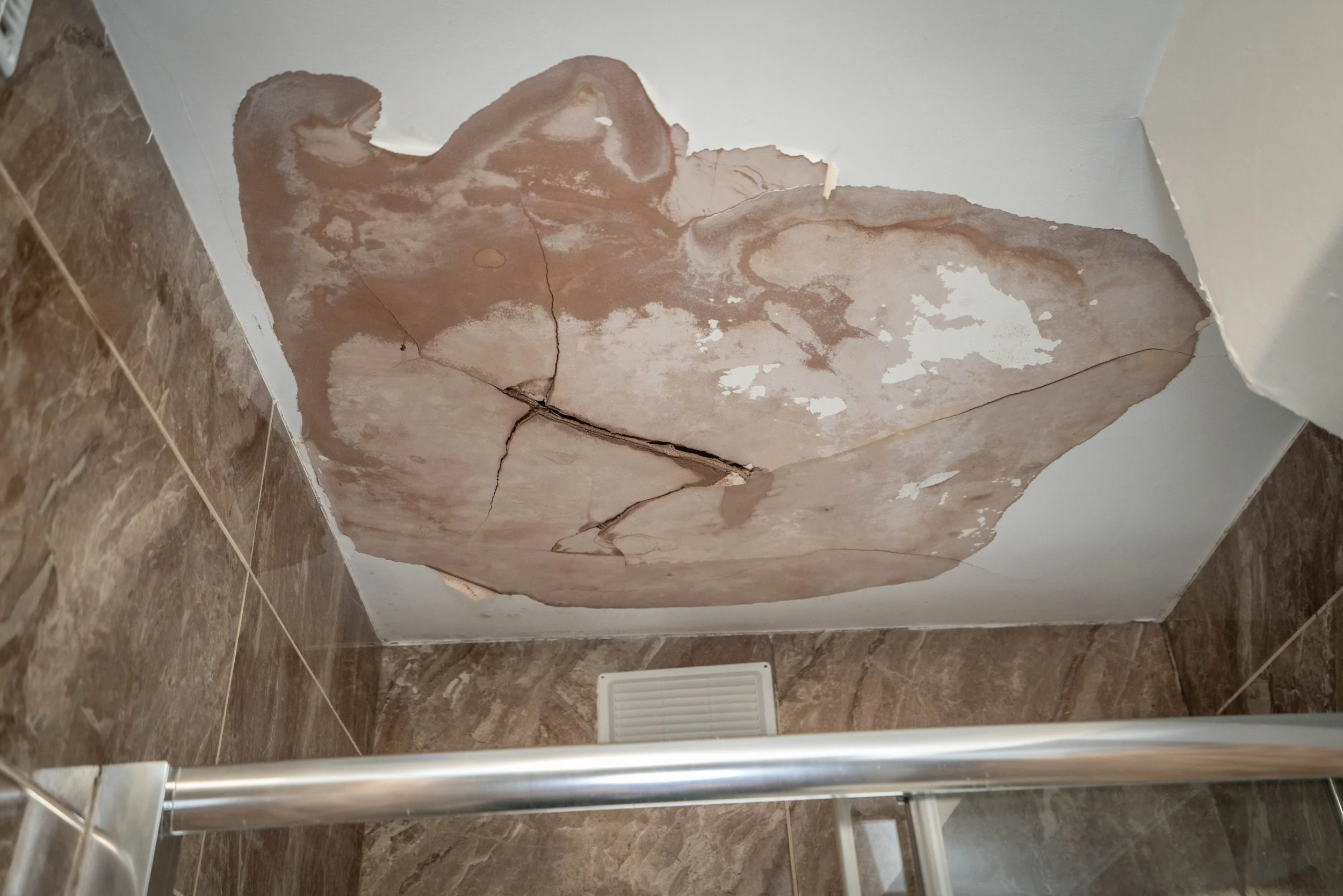 A bathroom corner with cracked and peeling wall paint or plaster, exposing underlying layers, and a brown-tiled floor.