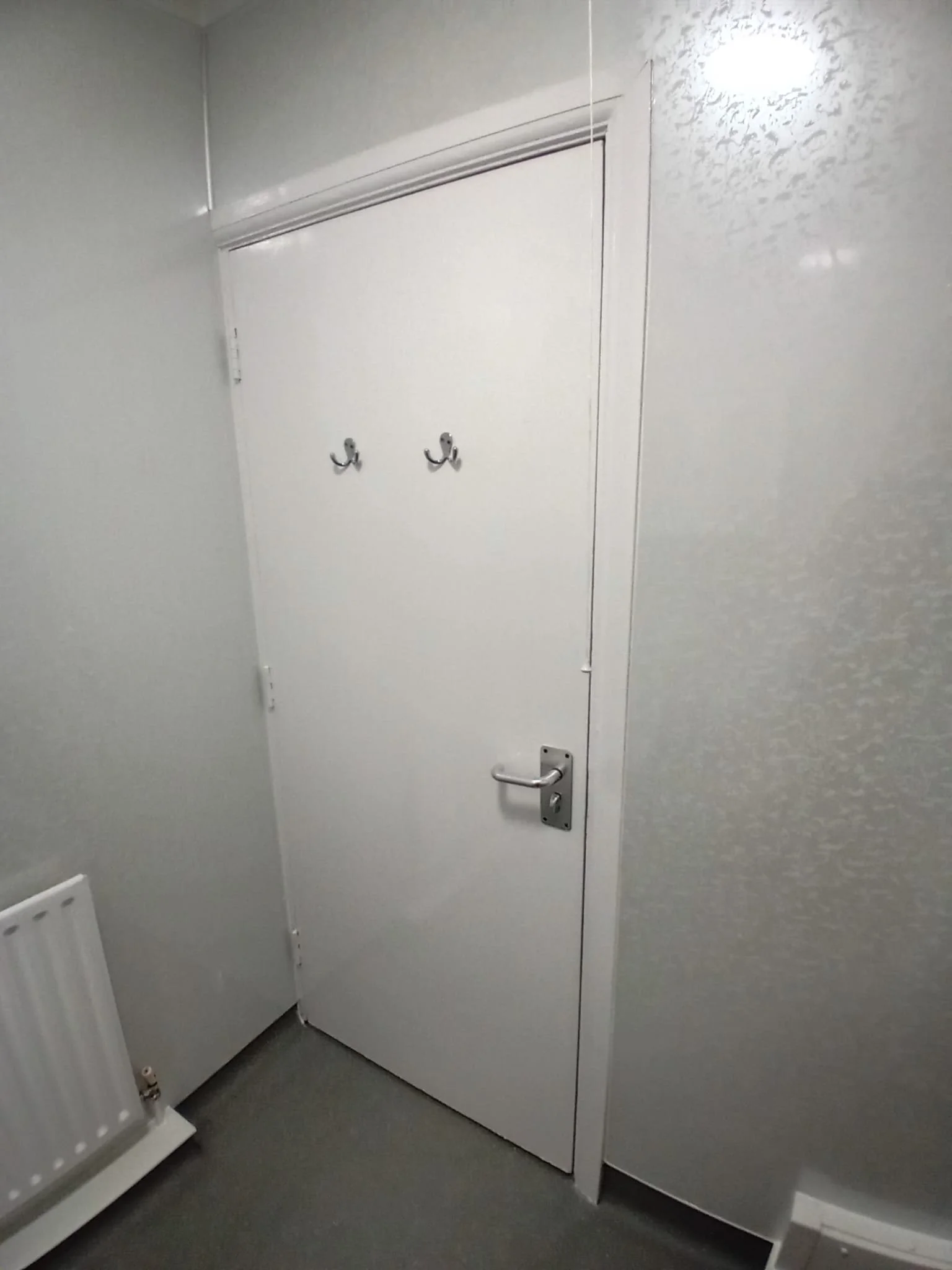 A white door with two hooks and a door handle in a small room, with a radiator visible on the left and a wall corner on the right.