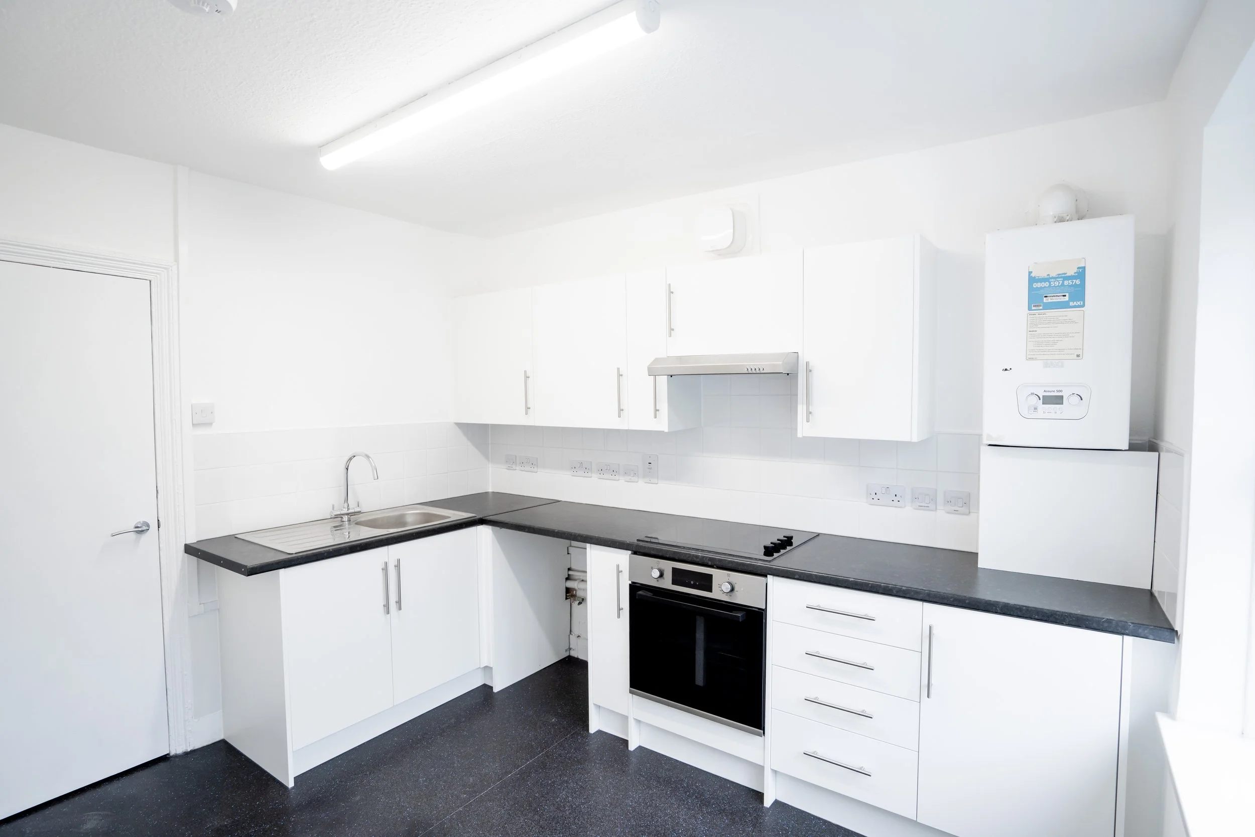 A clean, modern kitchen with white cabinets, black countertops, a stainless steel sink, a built-in oven, and a gas stove. There is a boiler mounted on the wall and several electrical outlets along the backsplash. The ceiling has a bright fluorescent 