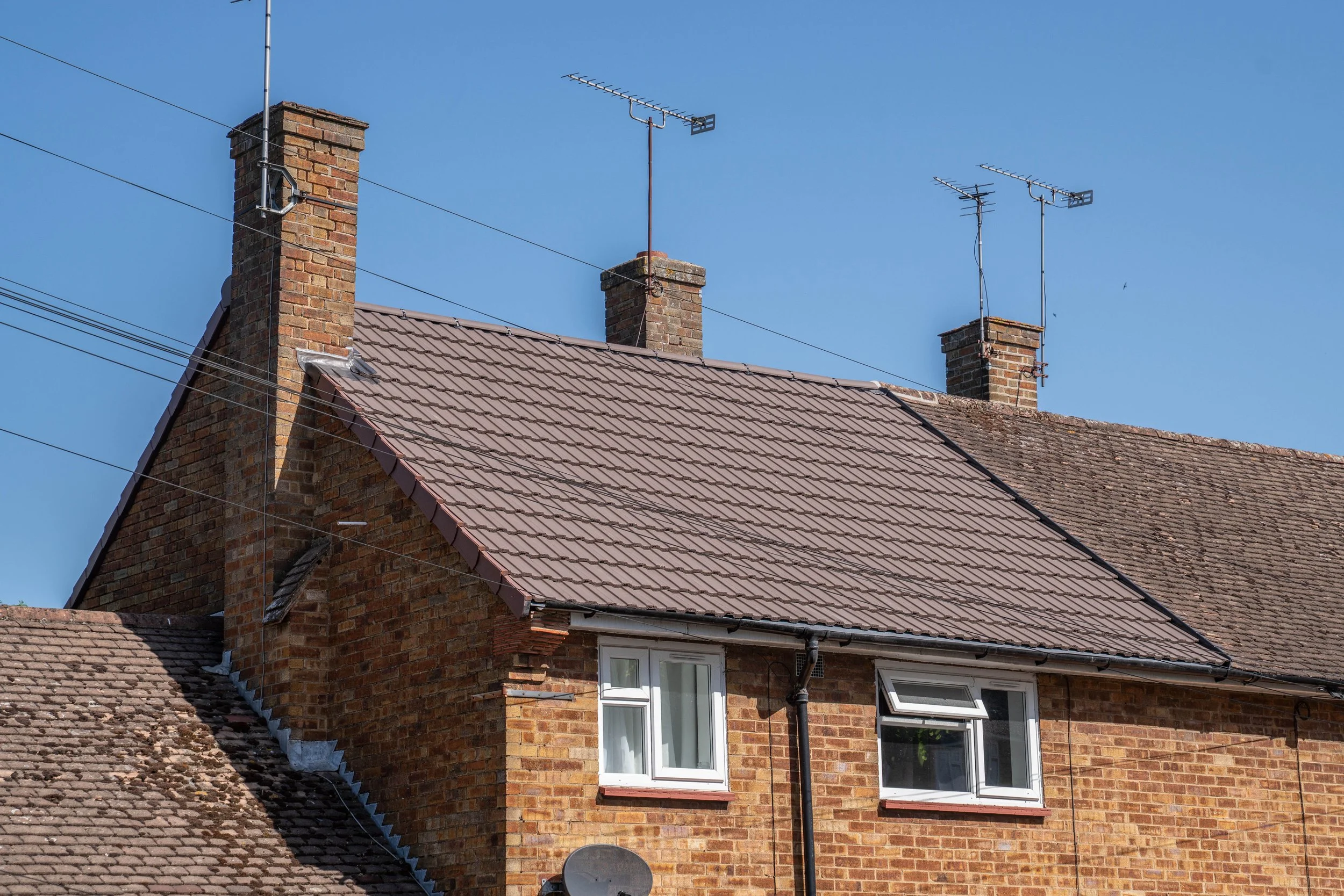 Brick house with multiple chimneys and television antennas on the roof under a clear blue sky.