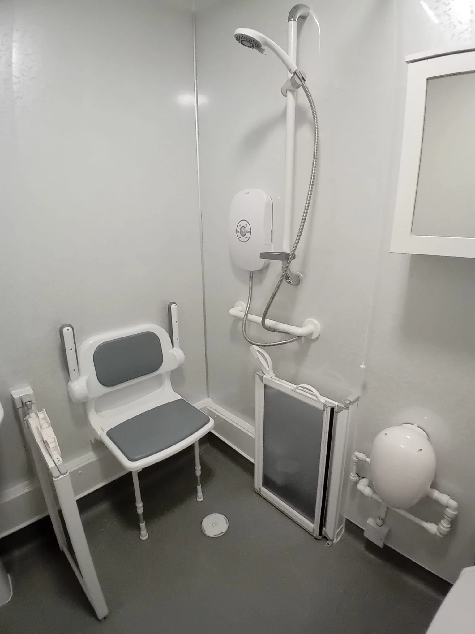 A compact shower room with a white shower head mounted on a wall, a white shower control panel, and a shower stool with white frame and gray cushion. The room has white walls and a gray floor, with a small circular drain on the floor.