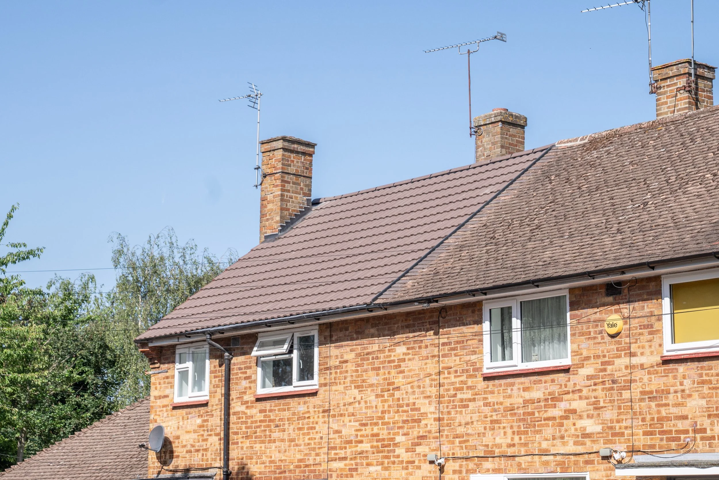 Close-up view of a brick house with a tiled roof, two chimneys, and two TV antennas on the roof. Part of a window with a satellite dish below is visible at the bottom.