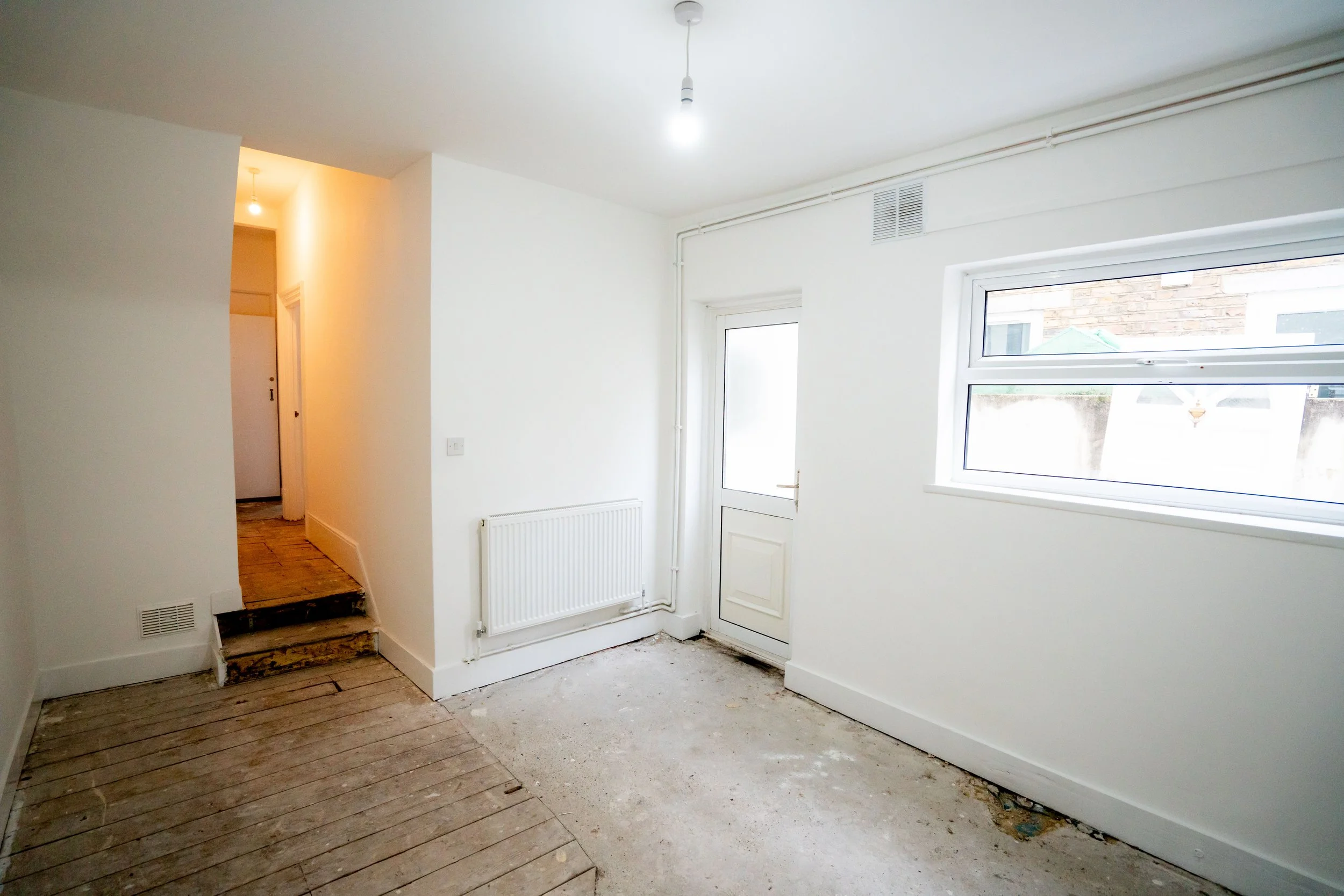 An unfinished room with a mix of new white walls and stripped wooden flooring, featuring a window and a door leading outside, with a section of floor under renovation.