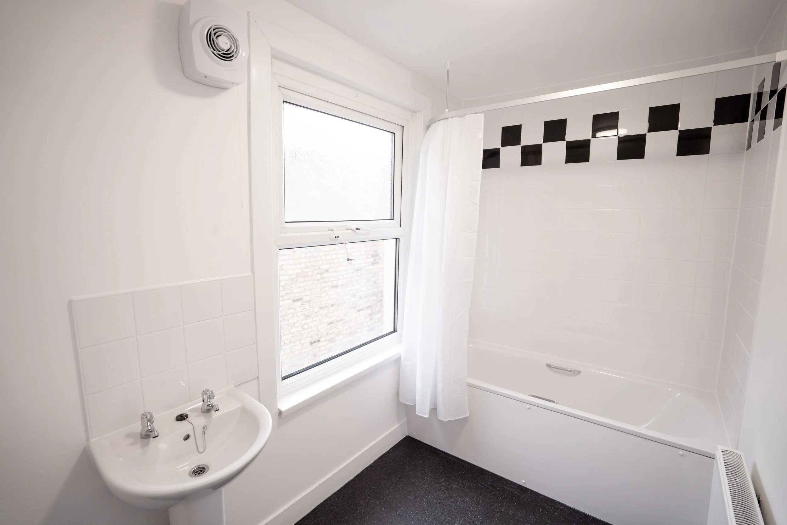 A bathroom featuring a white corner bathtub with a white shower curtain, a white sink with dual faucets, a window with a white frame, white tiled walls with a black checkerboard border, a wall-mounted vent, and a heather on a dark speckled floor.