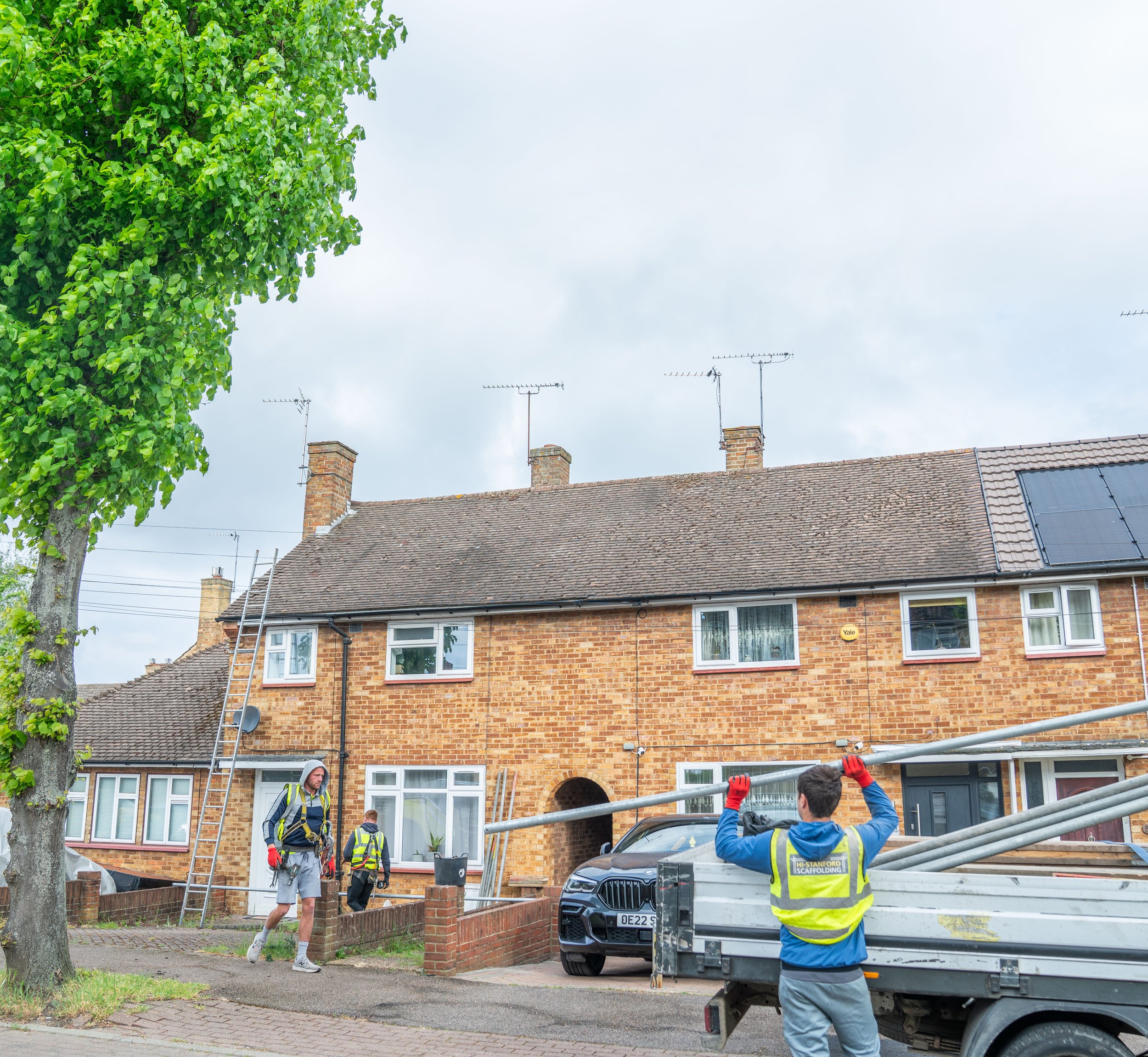Workers installing scaffolding in front of a brick house on a residential street. One worker is wearing a high-visibility vest with the words 'HIGH-LEVEL SCAFFOLDING' on the back. There are trees and television antennas visible on the rooftops.