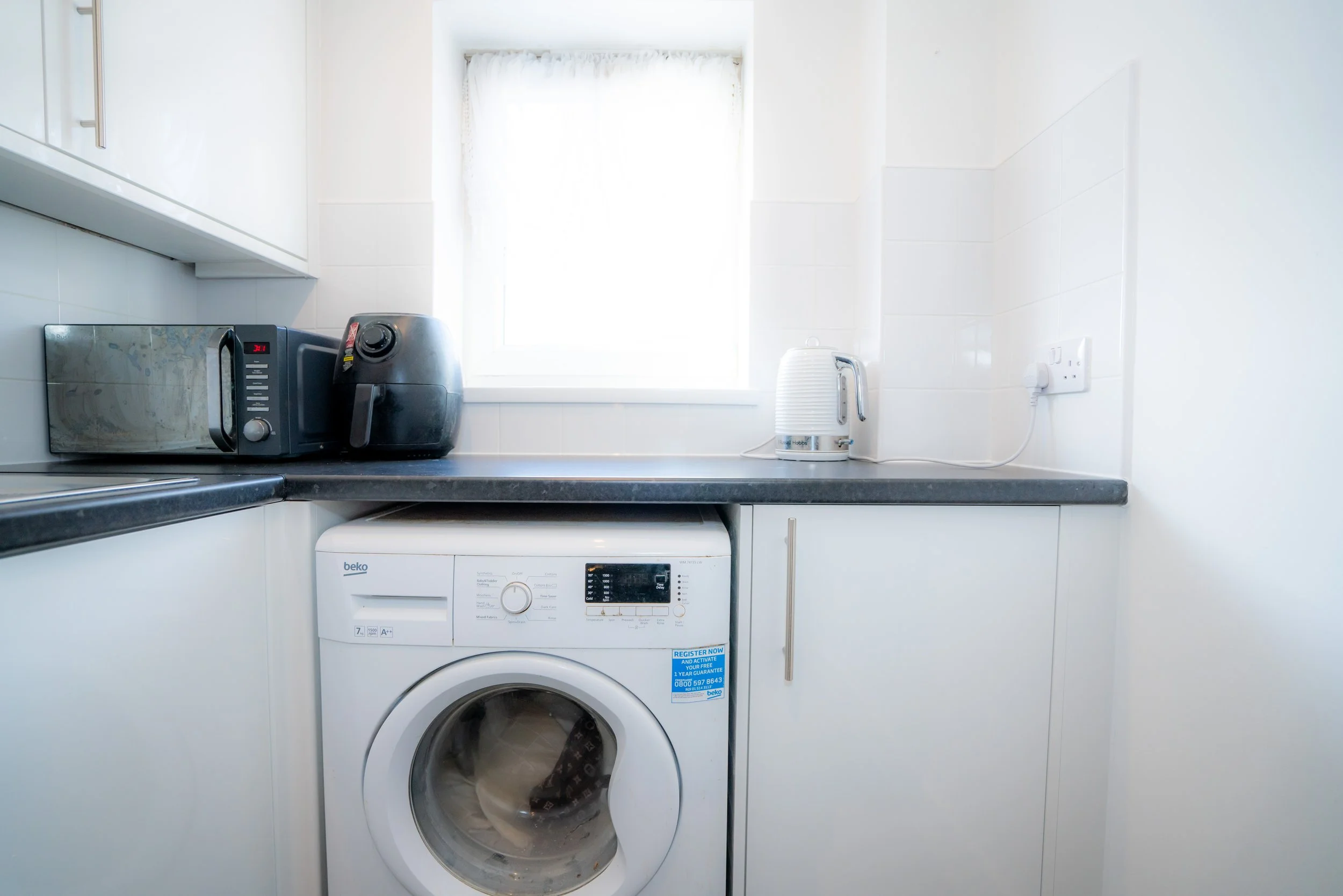 A laundry area with a washing machine, a microwave, an air fryer, an electric kettle, and a window with white curtains.