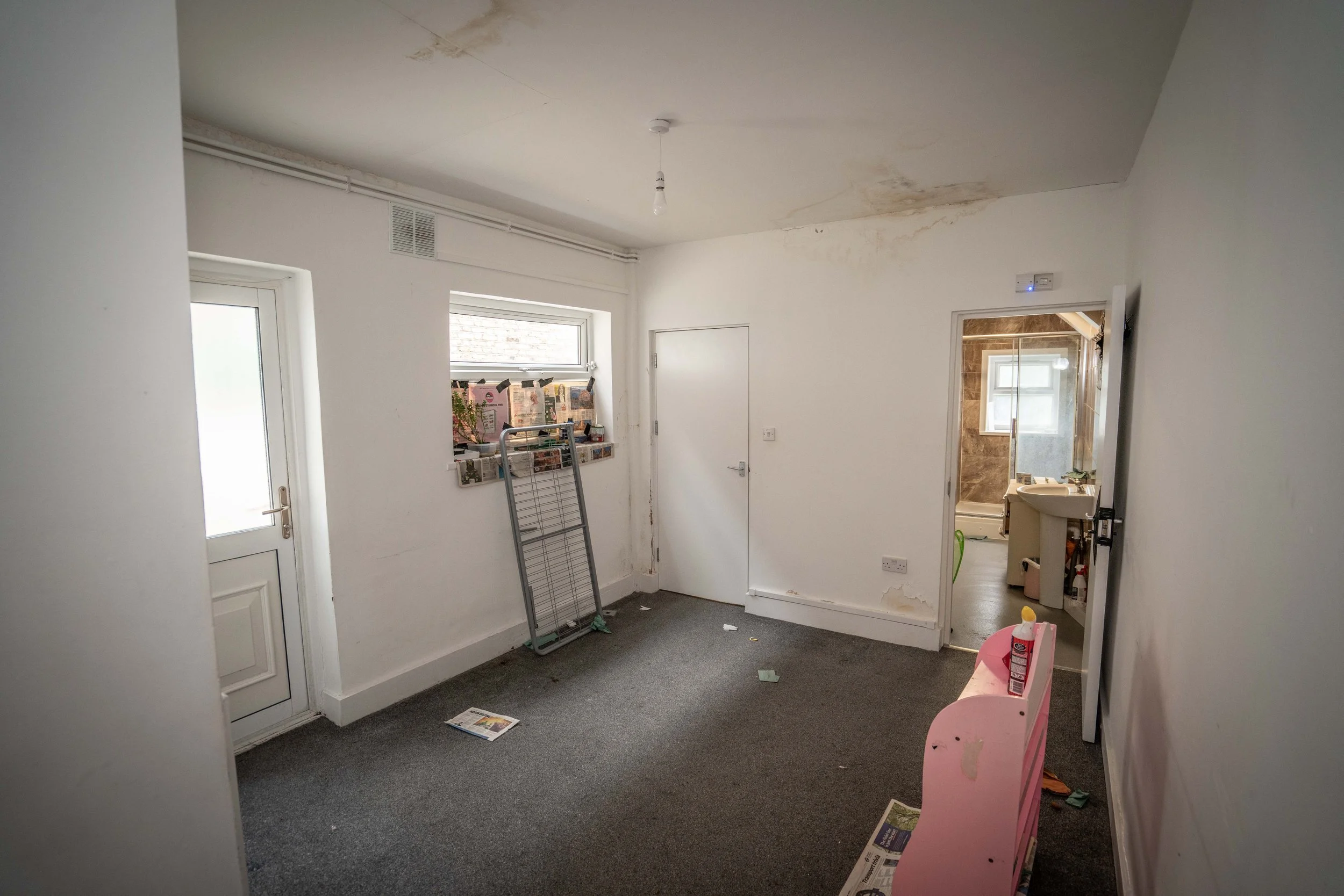 Room with white walls and grey carpet, showing water stains on ceiling and wall, a door leading outside, a window with newspapers on the windowsill, a small sink area, and a pink children's chair.