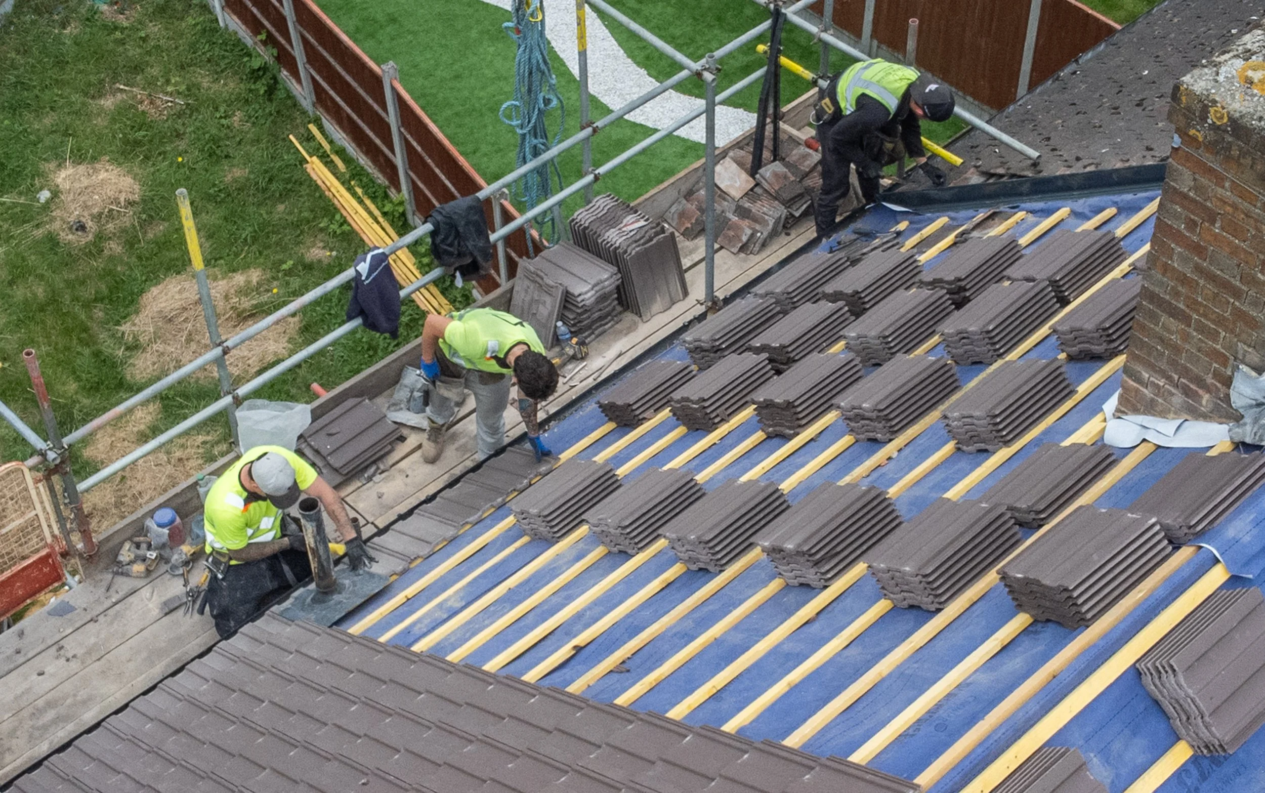 Construction workers installing roof tiles on a building, with scaffolding and construction materials around.
