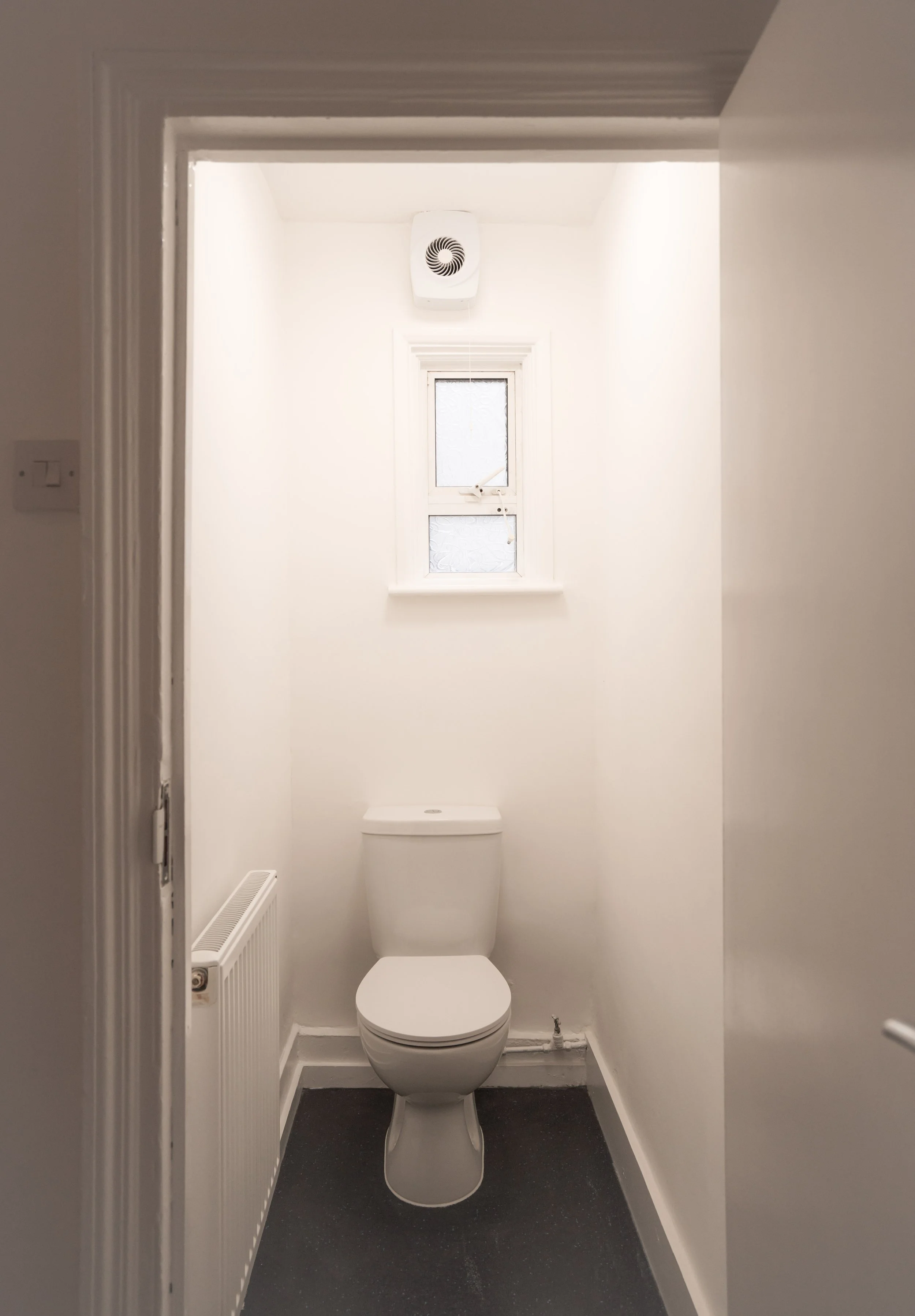 A small, narrow bathroom with a white toilet, a small window above, a white vent on the ceiling, a radiator on the left wall, and dark flooring.