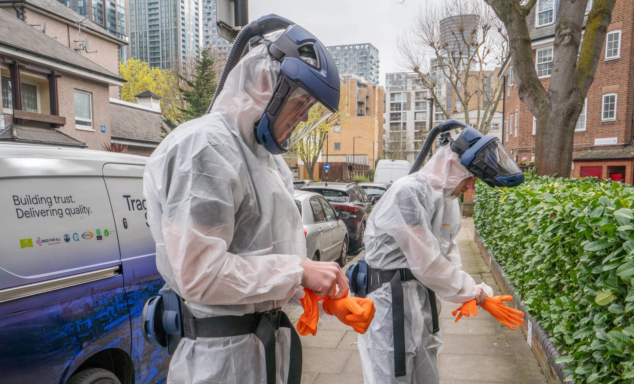 Two workers dressed in white hazmat suits, wearing gloves, and face shields, are inspecting a row of green bushes on a city sidewalk. A vehicle with a slogan on the side is parked nearby, and in the background are trees, residential buildings, and hi