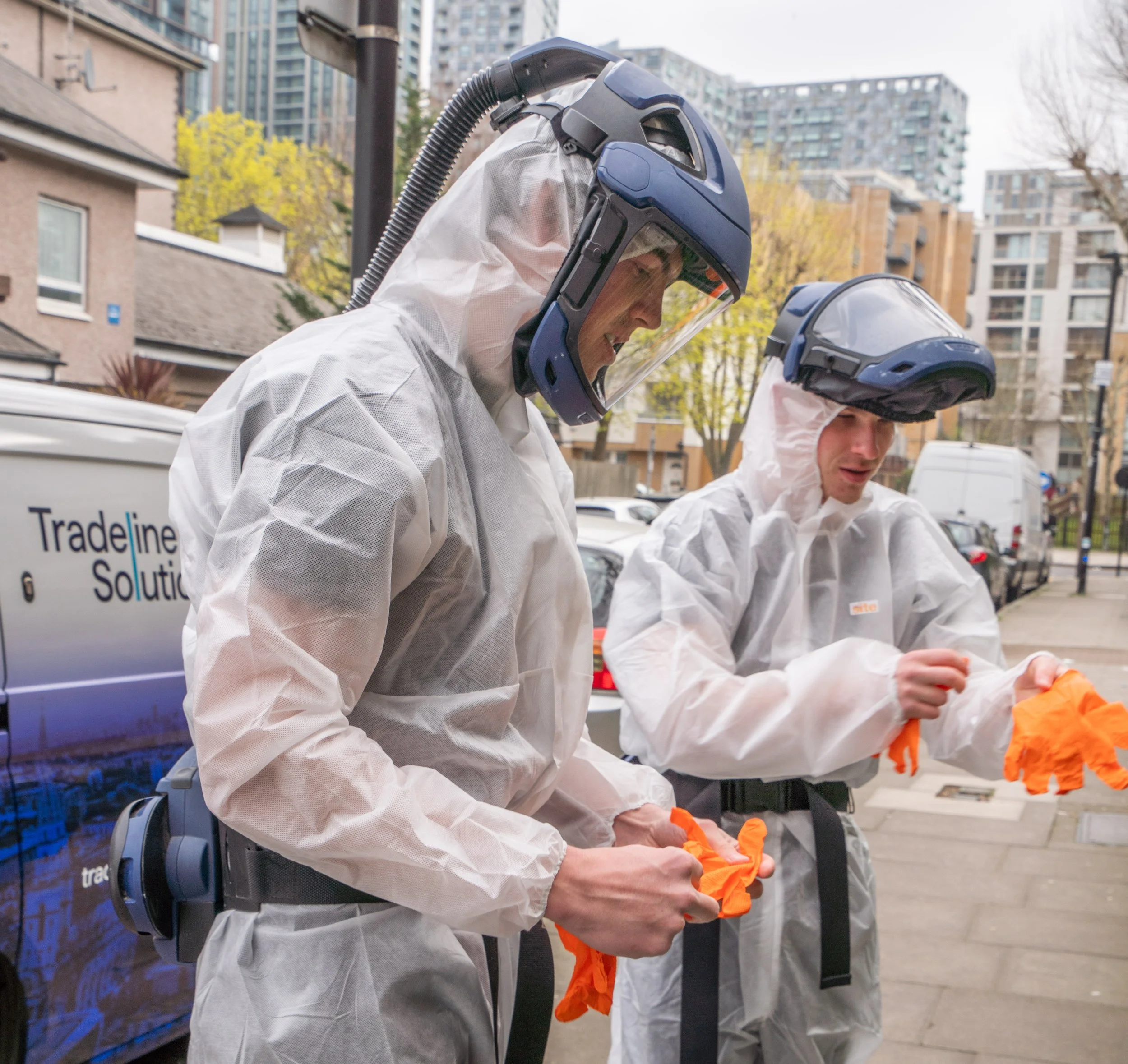 Two people wearing white protective suits, face shields, and helmets with respirators, standing outdoors in an urban area with high-rise buildings and trees in the background.