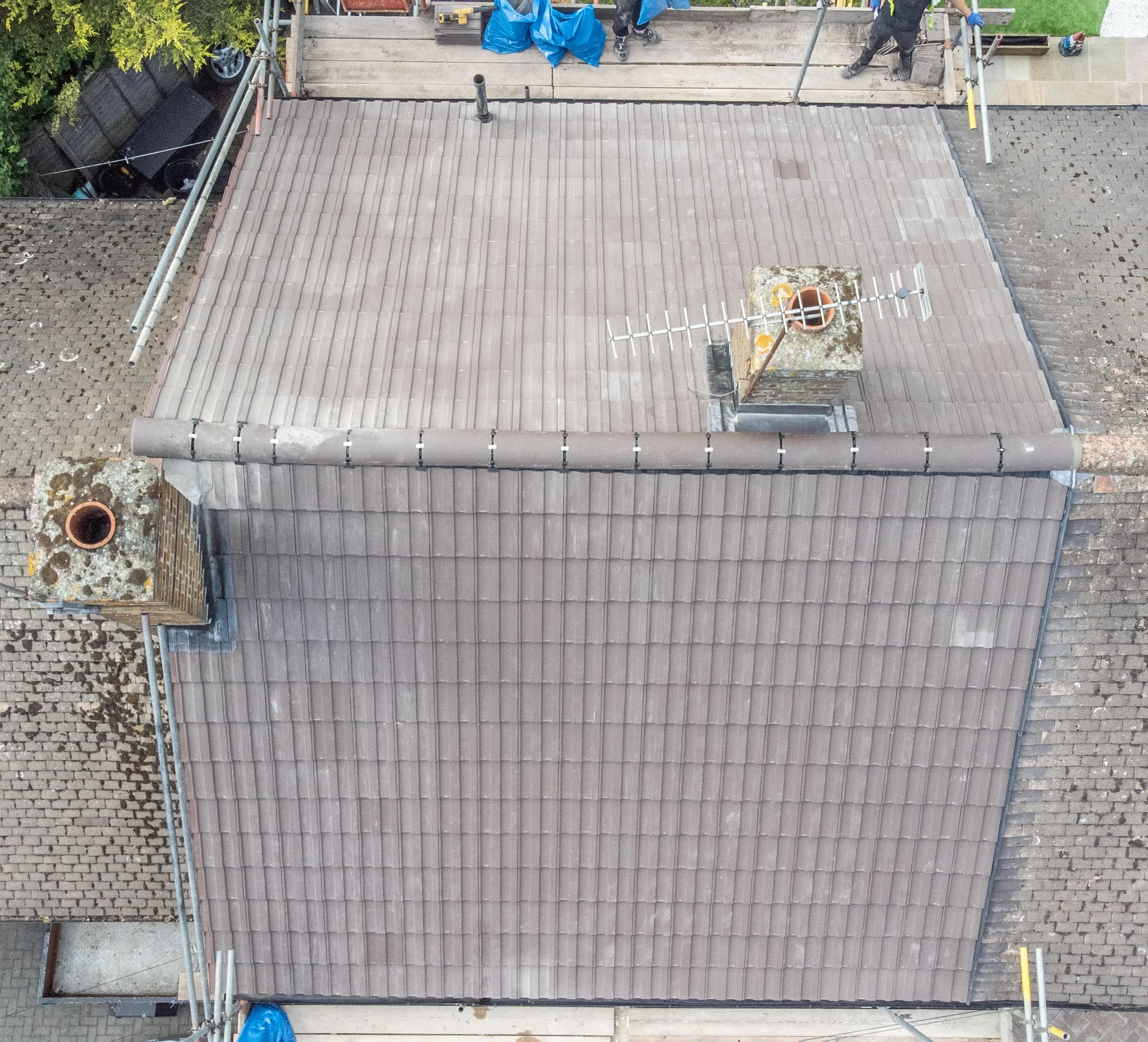 Aerial view of a roof undergoing construction or repair, with a section of new tiles and workers on a wooden platform nearby.