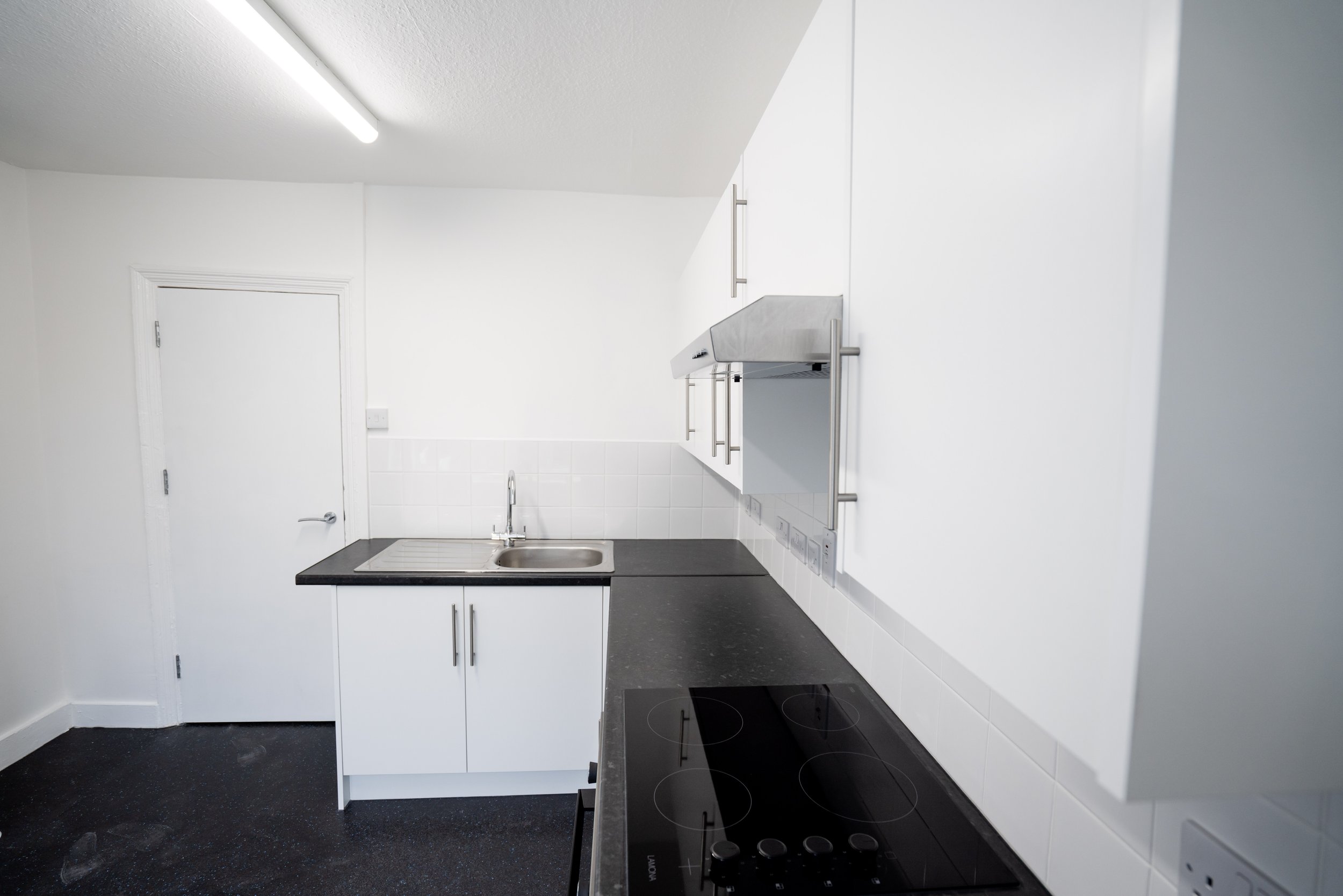 A small, white kitchen with white cabinets, black countertop, white tiled backsplash, stainless steel sink, and an electric cooktop, with a white door on the left and fluorescent ceiling light.
