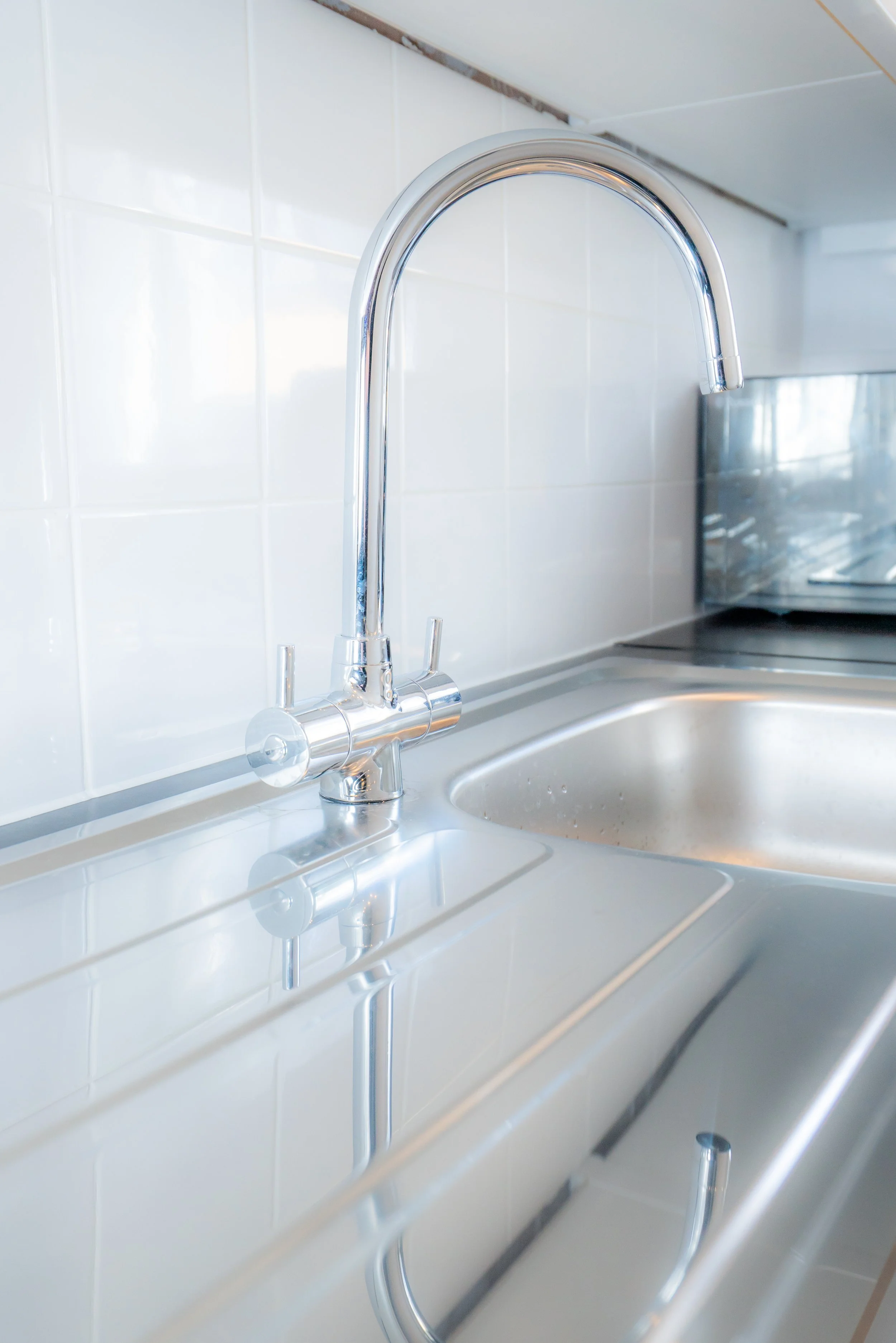 A kitchen sink with a chrome faucet, white tiled wall, and a stainless steel oven in the background.