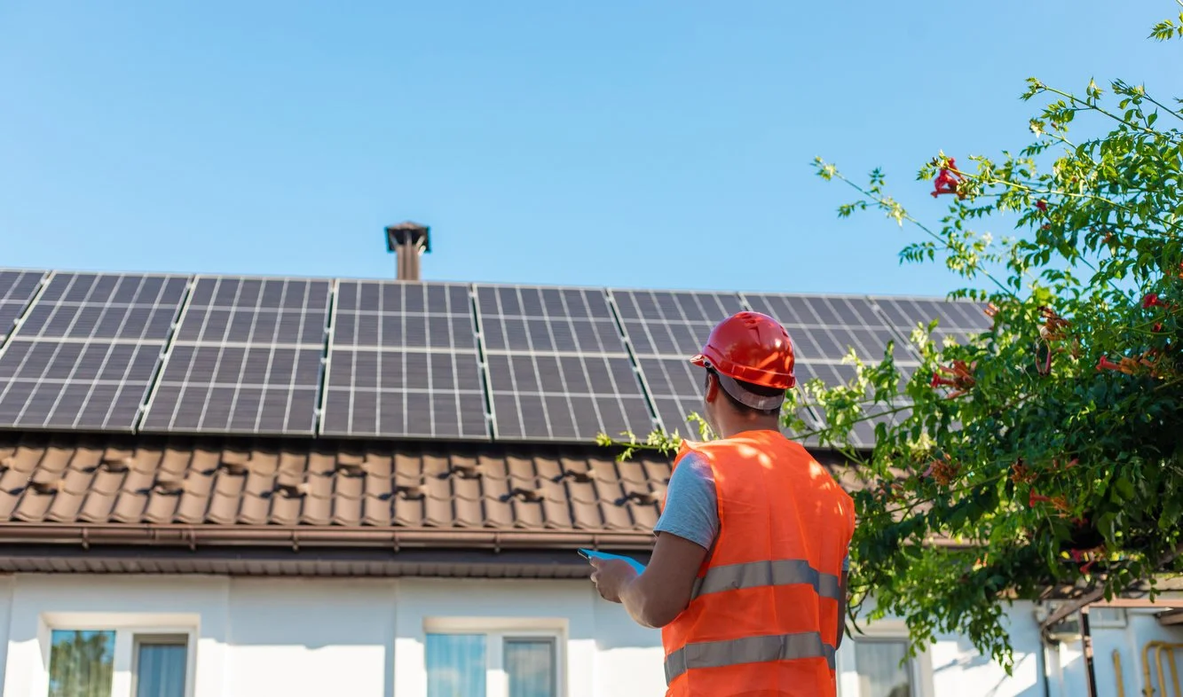 A worker in a safety vest and helmet standing outside a house with solar panels on the roof, looking at a phone.