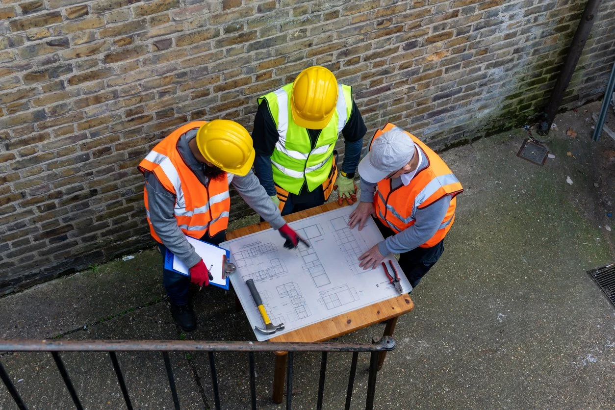 Three construction workers wearing safety vests and helmets gathered around a table with blueprints, discussing the plans outdoors against a brick wall.