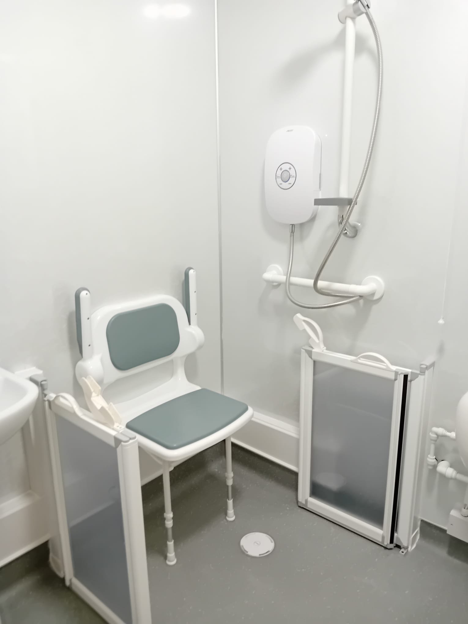 Hospital shower room with a foldable shower chair, a wall-mounted shower unit, a grab bar, and a full-length mirror.