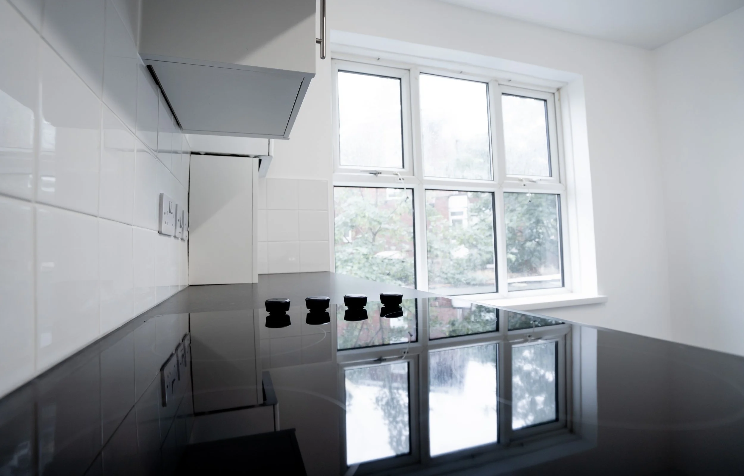 A modern kitchen with a black stove and a reflective black countertop, white tiled backsplash, and large windows showing trees outside.